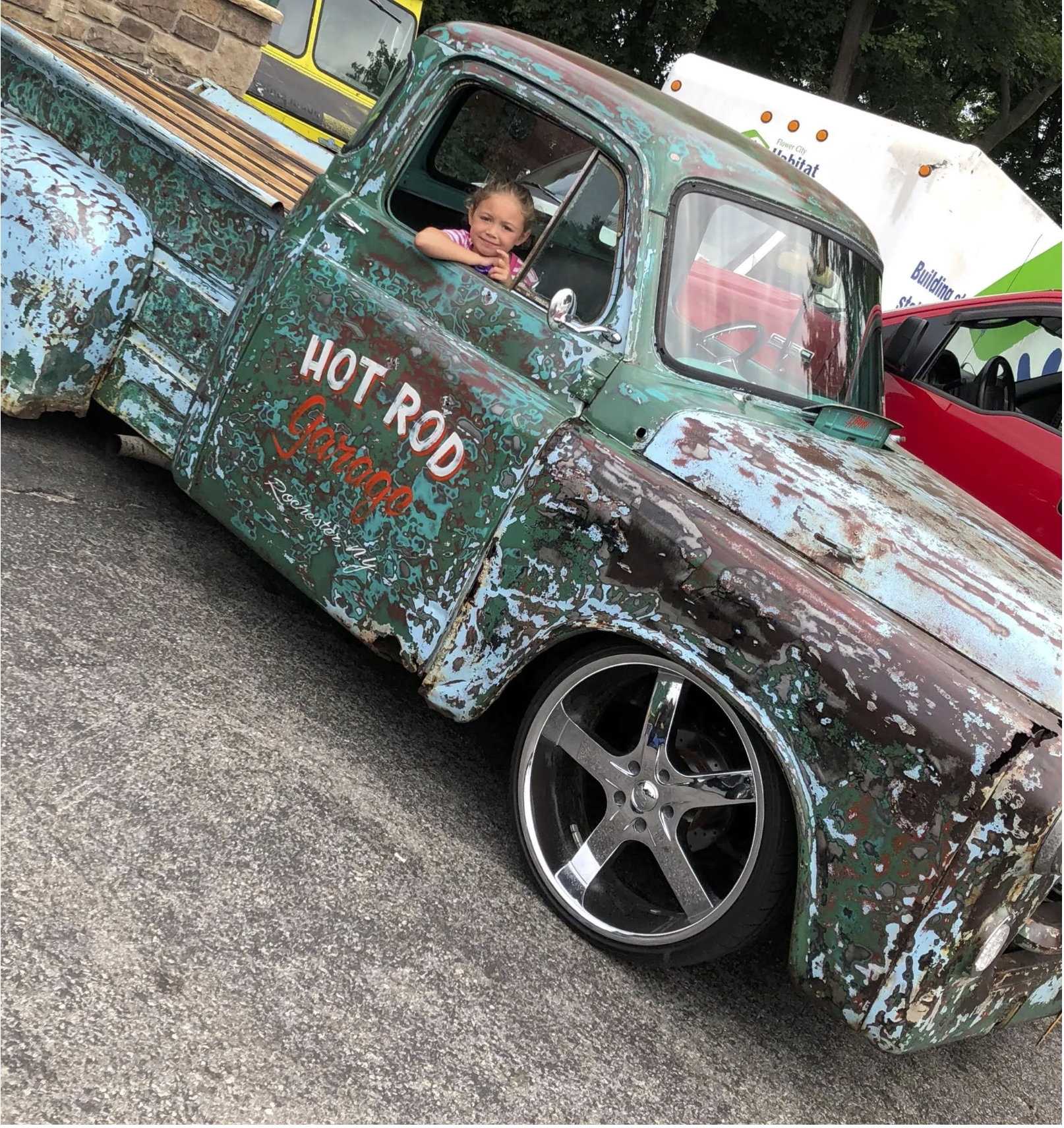A young girl with blonde hair, wearing a pink and white striped shirt, sits inside an old, rusted pickup truck with a colorful, weathered paint job. The truck has a sign on the door that reads 'HOT ROD Something.' The truck is parked on a street with other vehicles and trees in the background.