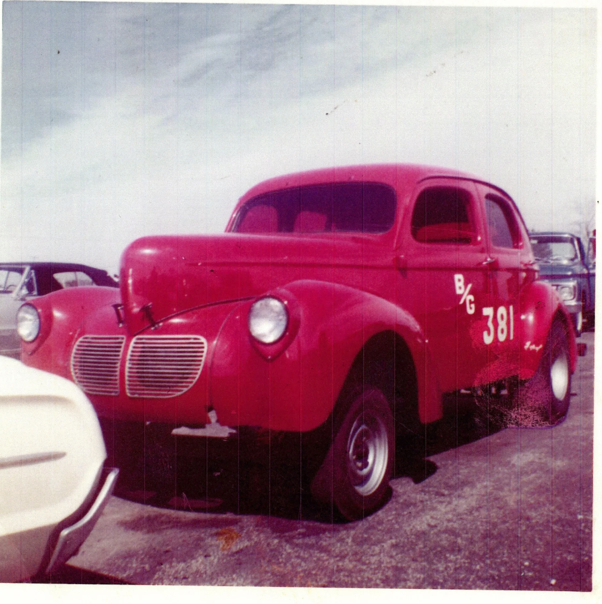 Vintage red race car with the number 381 and B/G painted on the side, parked among other classic cars in an outdoor lot.