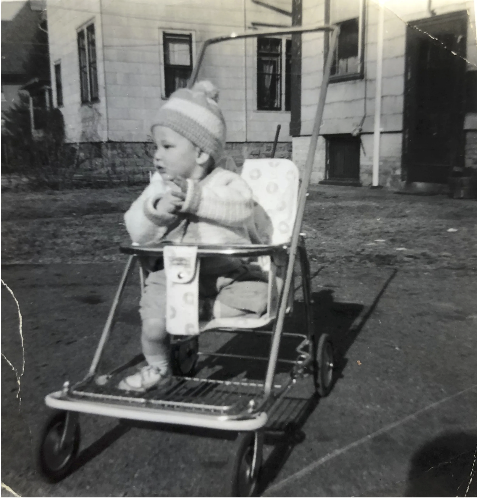 A young child in a high chair outdoors, sitting on pavement in front of residential houses. The child is wearing a knit hat and sweater, and appears to be holding a small object or food. There is a glass or cup on the tray of the high chair.