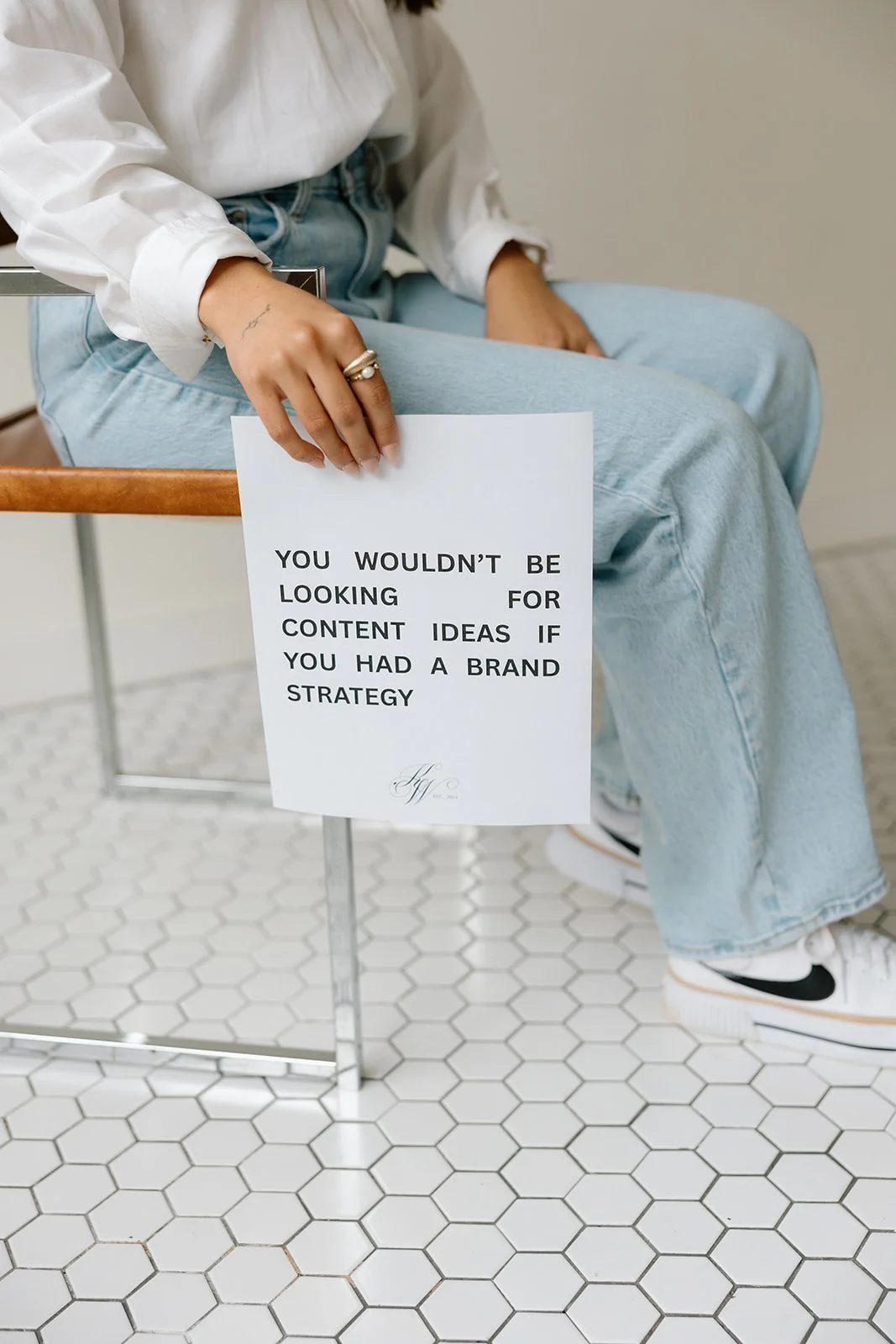 Person sitting on chair holding a sign with text about brand strategy, over hexagonal tile floor.
