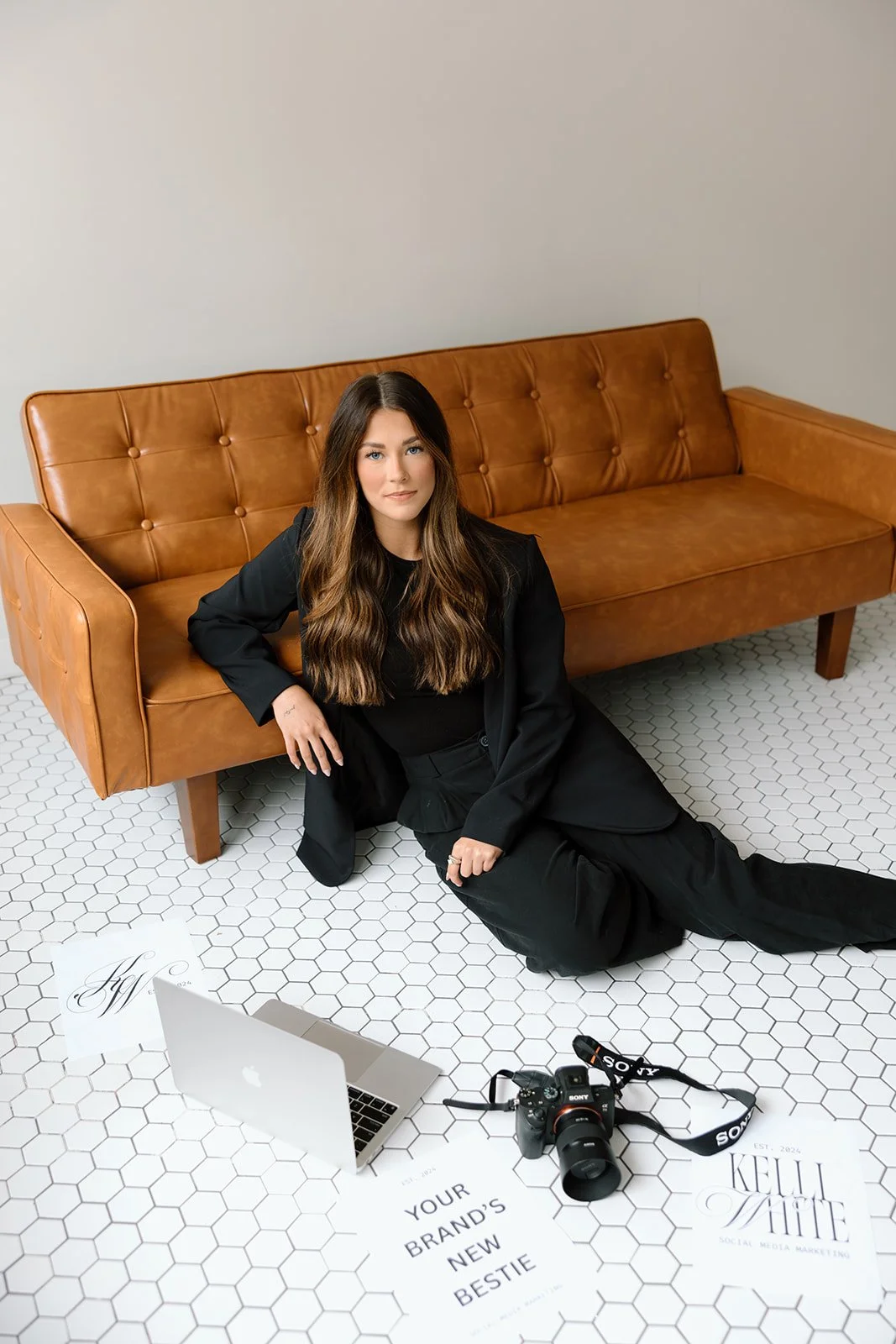 Woman sitting on the floor next to a brown leather couch, with a laptop, camera, and papers labeled "Your Brand's New Bestie" and "Kelli White Social Media Marketing" on a hexagon tile floor.