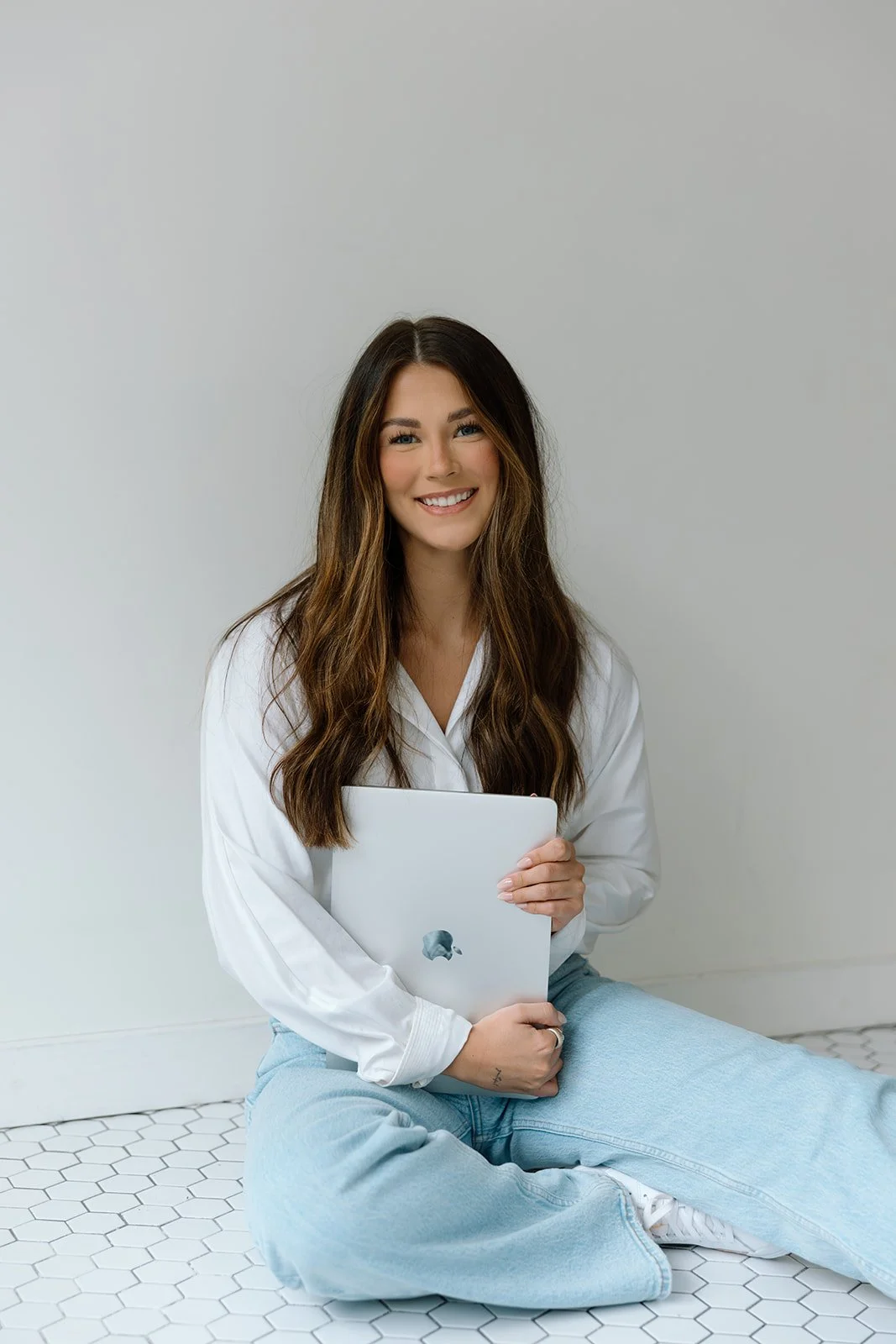 Woman sitting on a hexagon-tiled floor, holding a laptop, wearing a white shirt and blue jeans, smiling.