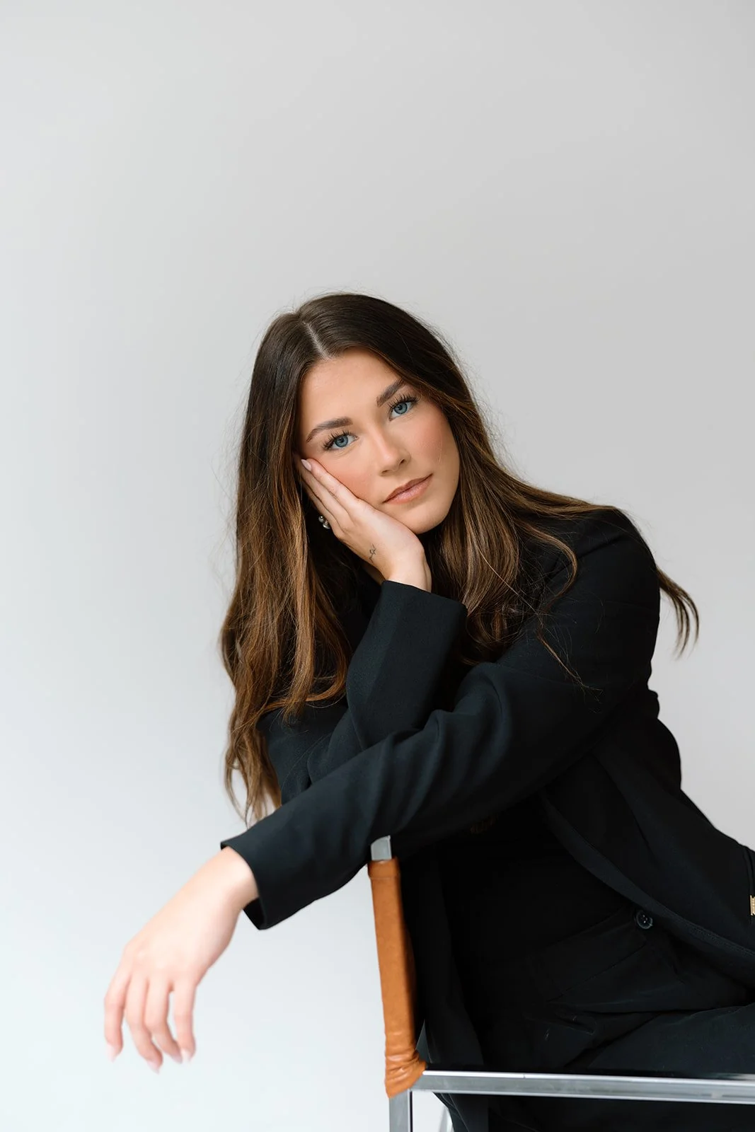 A woman with long brown hair and blue eyes sitting on a chair, resting her face on her hand, wearing a black blazer, in front of a plain white background.