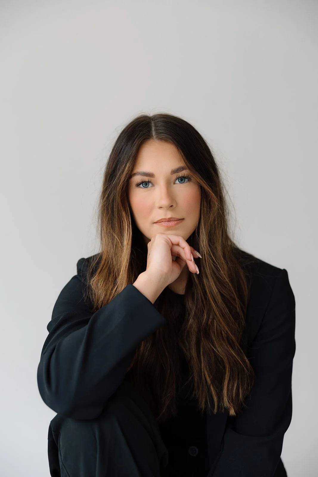 Woman with long brown hair wearing a black outfit, sitting and resting her chin on her hand, against a plain background.