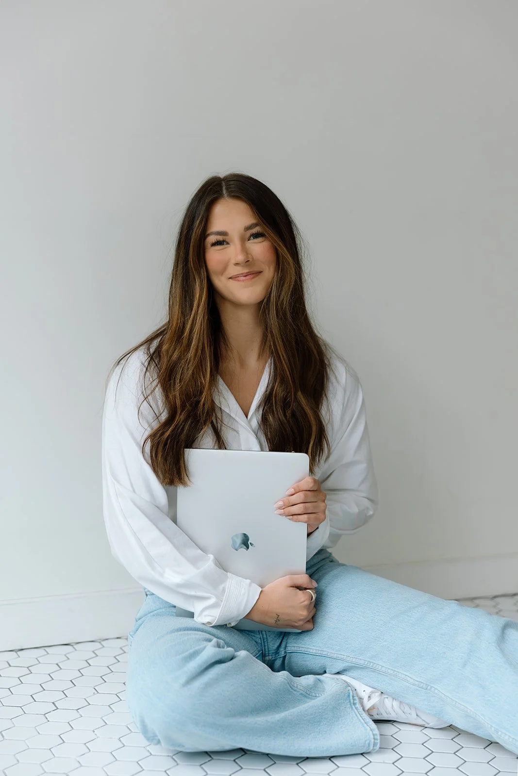 Woman in white shirt holding a laptop while sitting on a tiled floor.