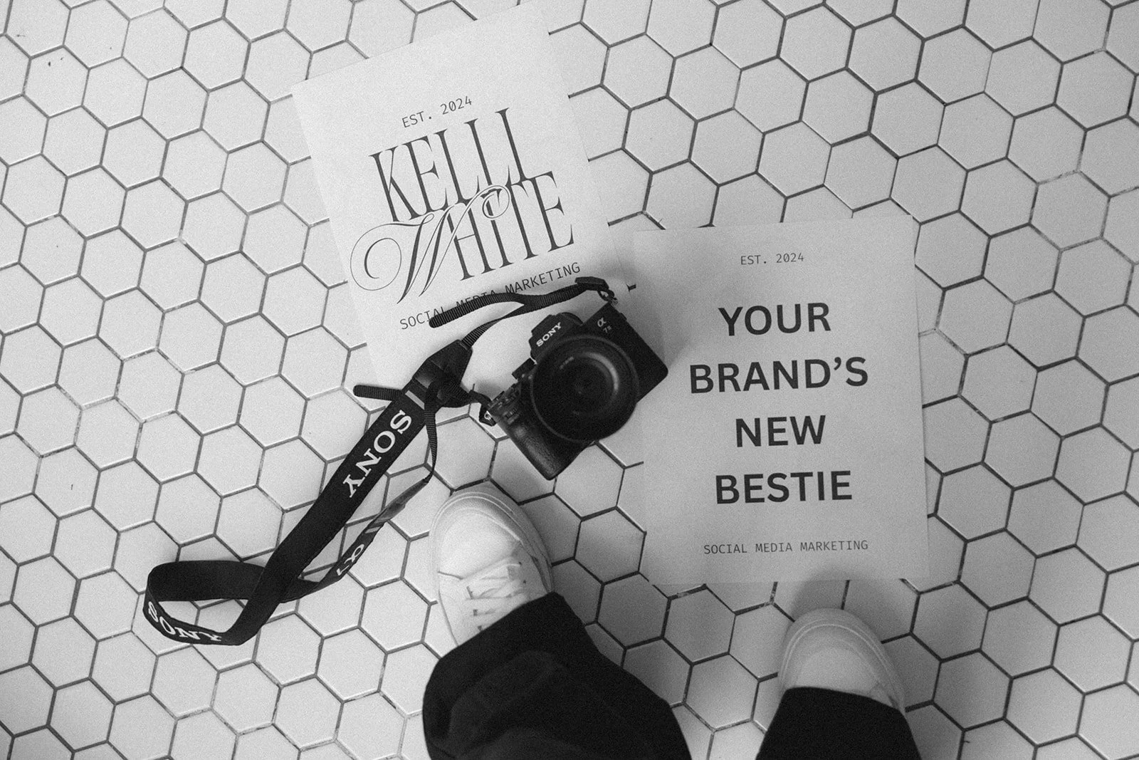 Black and white photo of a Sony camera on a hexagonal tile floor, with papers reading "Kelli White Social Media Marketing" and "Your Brand's New Bestie." Feet wearing white sneakers are partially visible.