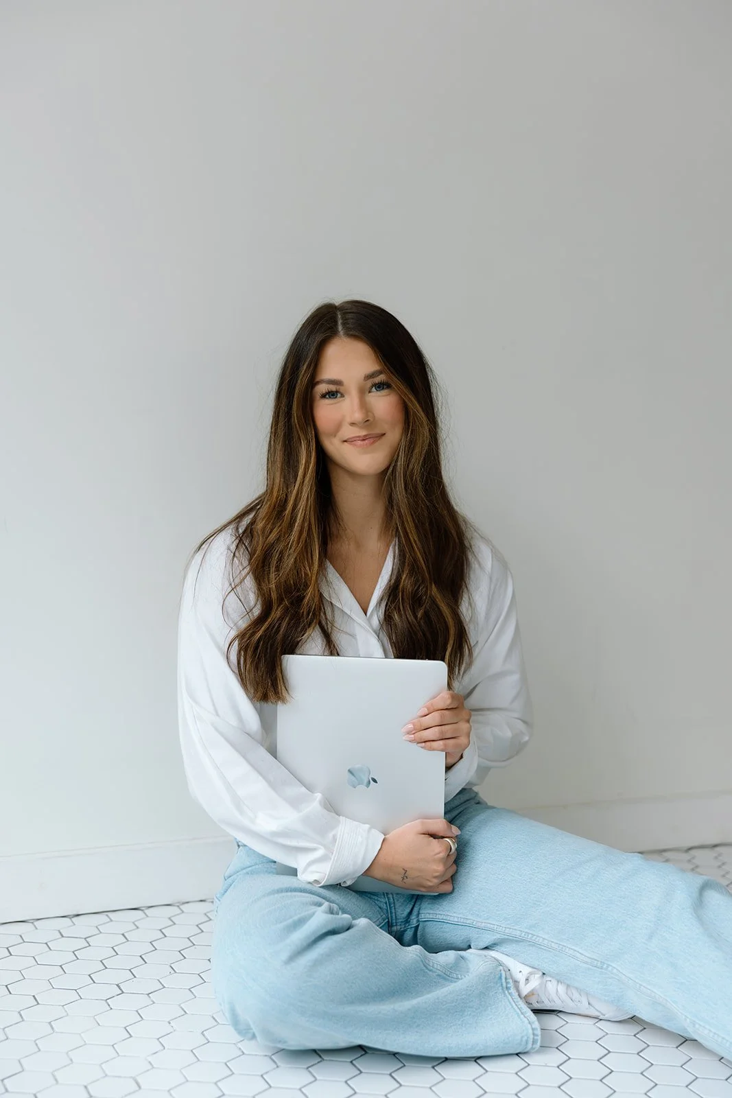 A young woman with long wavy brown hair sitting on a white tiled floor, holding a closed silver laptop, smiling at the camera, wearing a white blouse and light blue jeans, against a plain light grey wall.