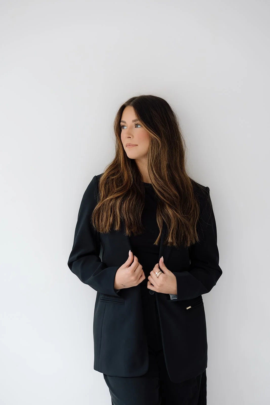 A woman with long, wavy brown hair and fair skin standing against a plain white background. She is wearing a black blazer and black top, and is looking off to the side.