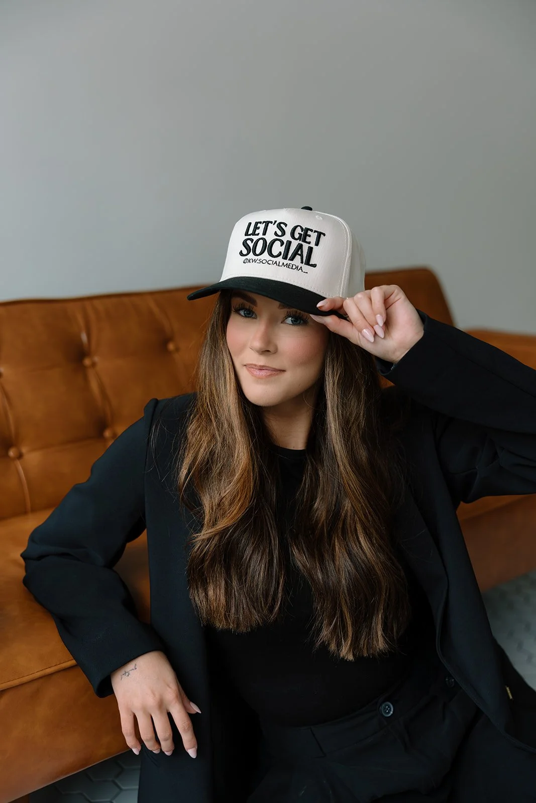 A woman with long, wavy brown hair sitting on a brown leather couch, wearing a black jacket and top, and a white and black baseball cap that says 'Let's Get Social'.
