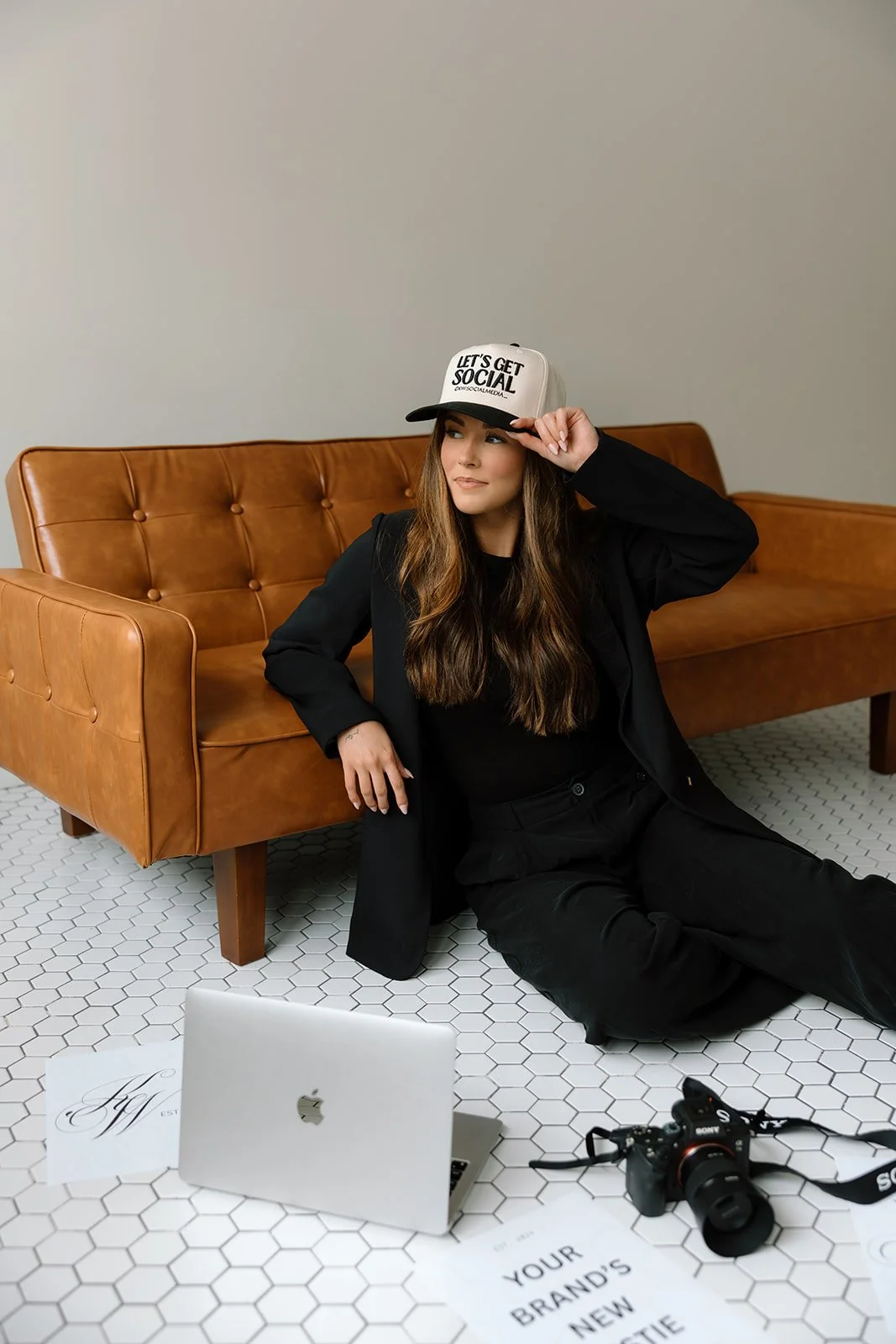 Person sitting on floor in front of a brown leather sofa, wearing a black outfit and a cap with "Let's Get Social" text. Nearby are a laptop, camera, and some papers on a tiled floor.
