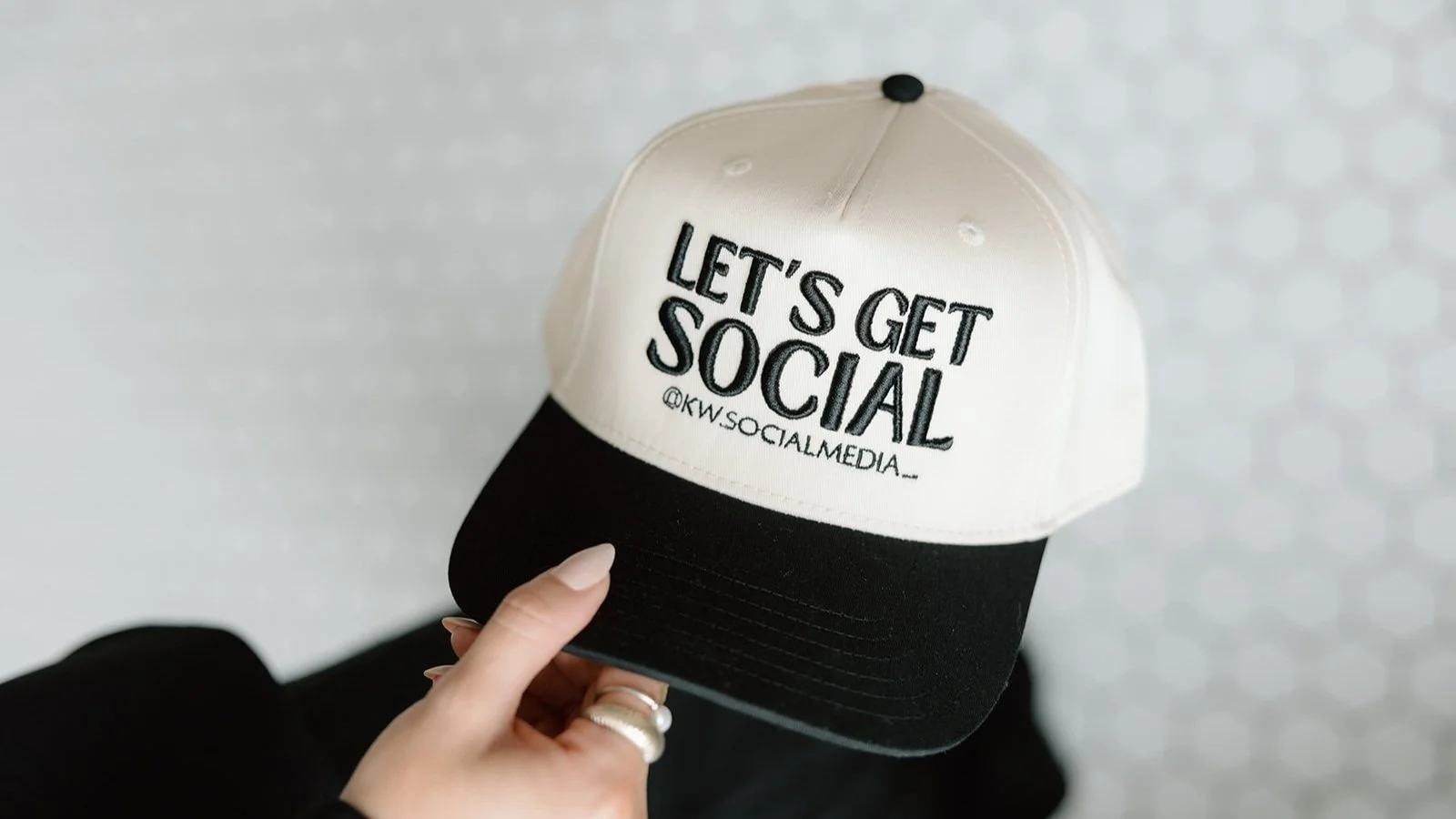 A beige and black baseball cap with the text "LET'S GET SOCIAL @KW.SOCIALMEDIA_" held by a hand with rings.
