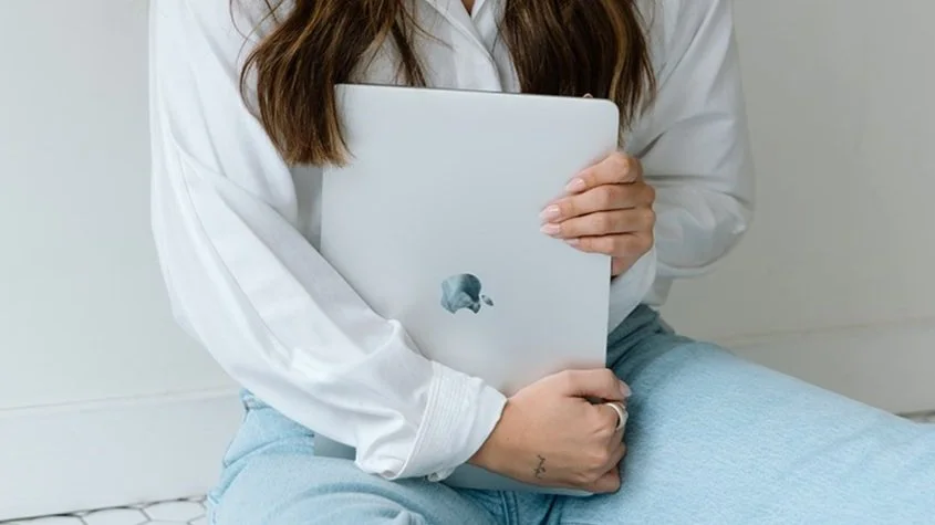 Person holding a closed laptop with an apple logo, wearing a white shirt and light blue jeans.
