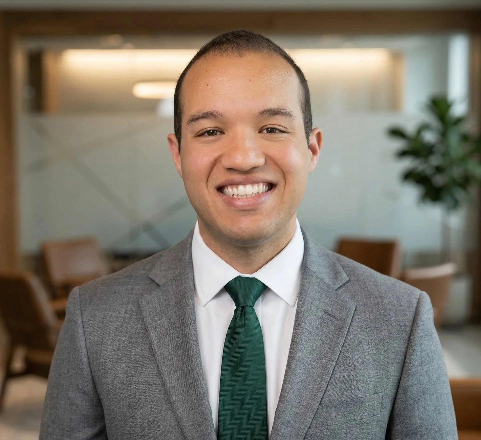 A smiling man in a gray suit, white shirt, and green tie, standing in a modern office with blurred background decor and seating.