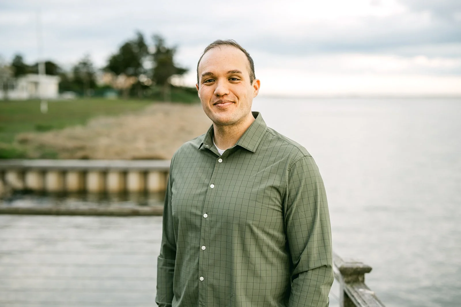 A man in a green checkered shirt standing by a waterfront with a cloudy sky in the background.