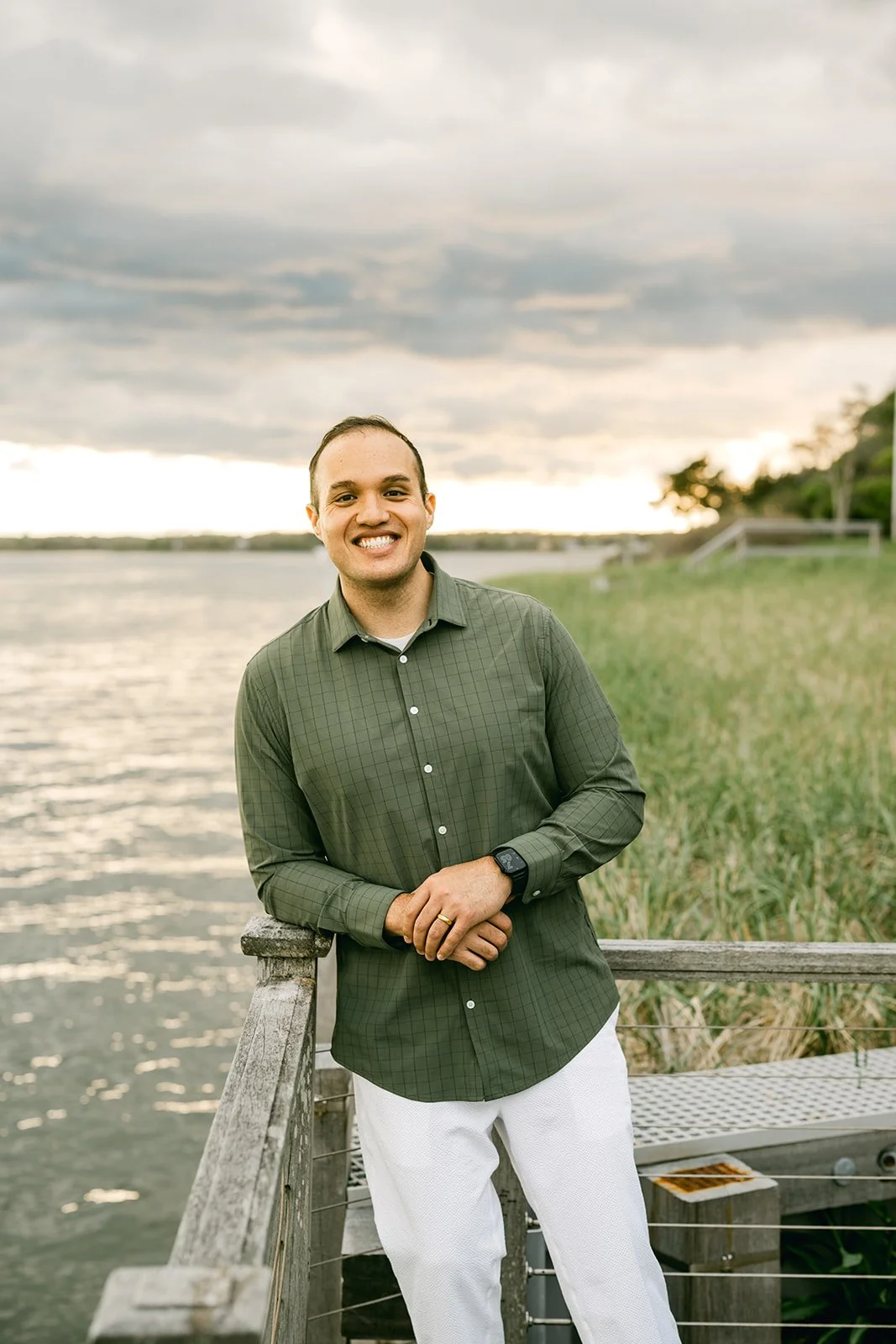A man smiling, wearing a green checkered shirt, standing on a dock near a body of water with a cloudy sky in the background.