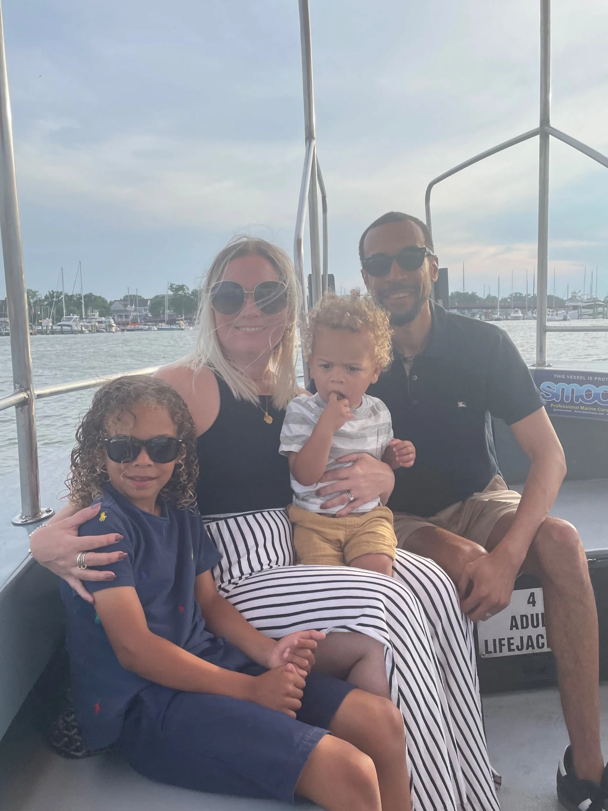 A family of five on a boat near a marina, with water and boats in the background. The woman is smiling, wearing sunglasses, and has blonde hair. The man is sitting next to her, also wearing sunglasses. Two children are in the photo, one with curly hair and the other with straight hair, all smiling and enjoying the moment.