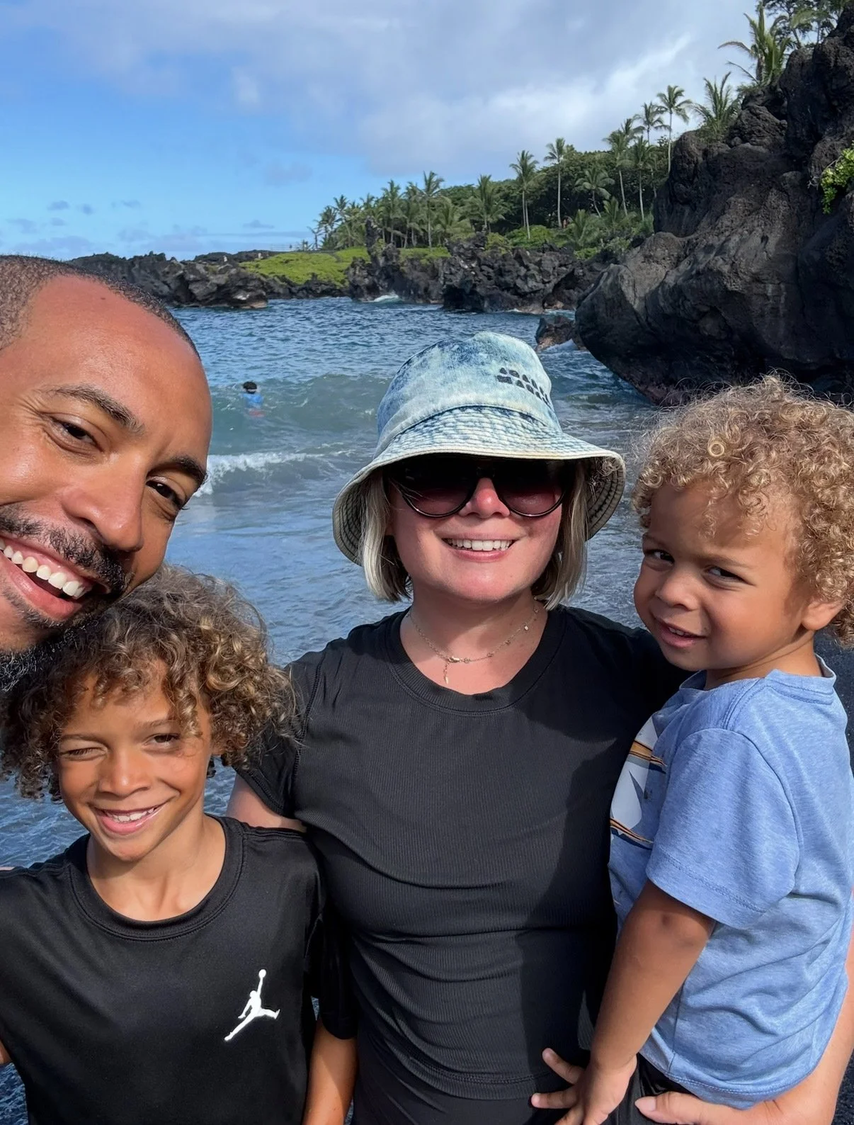 A group of five people smiling at the camera on a beach, with the ocean, rocks, and palm trees in the background. The group includes two adult women, one adult man, and two young boys, one with curly hair.