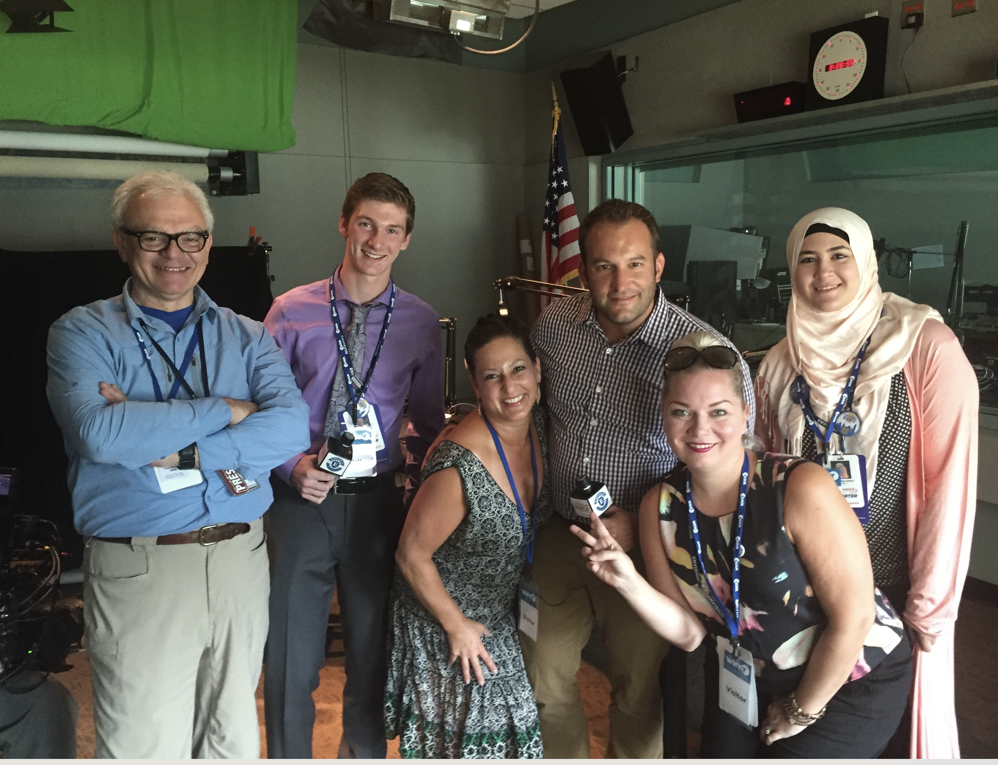 Group of eight diverse people smiling and posing for a photo in a broadcasting studio, some holding microphones, with a green screen, American flag, and broadcasting equipment visible in the background.