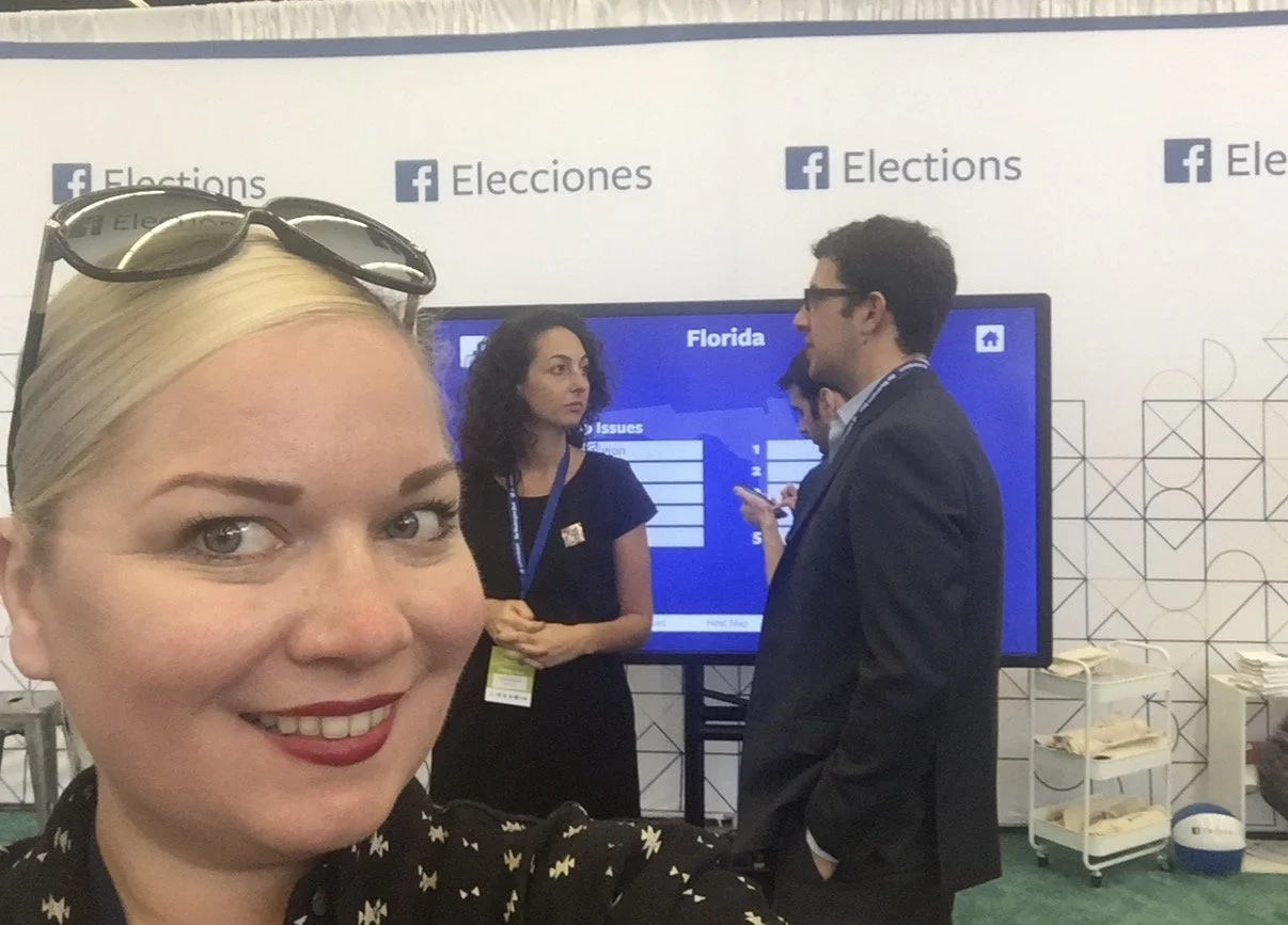 A woman with blonde hair, glasses on her head, and a black blouse with a star pattern taking a selfie at a voting or election event with several people and a large screen in the background.