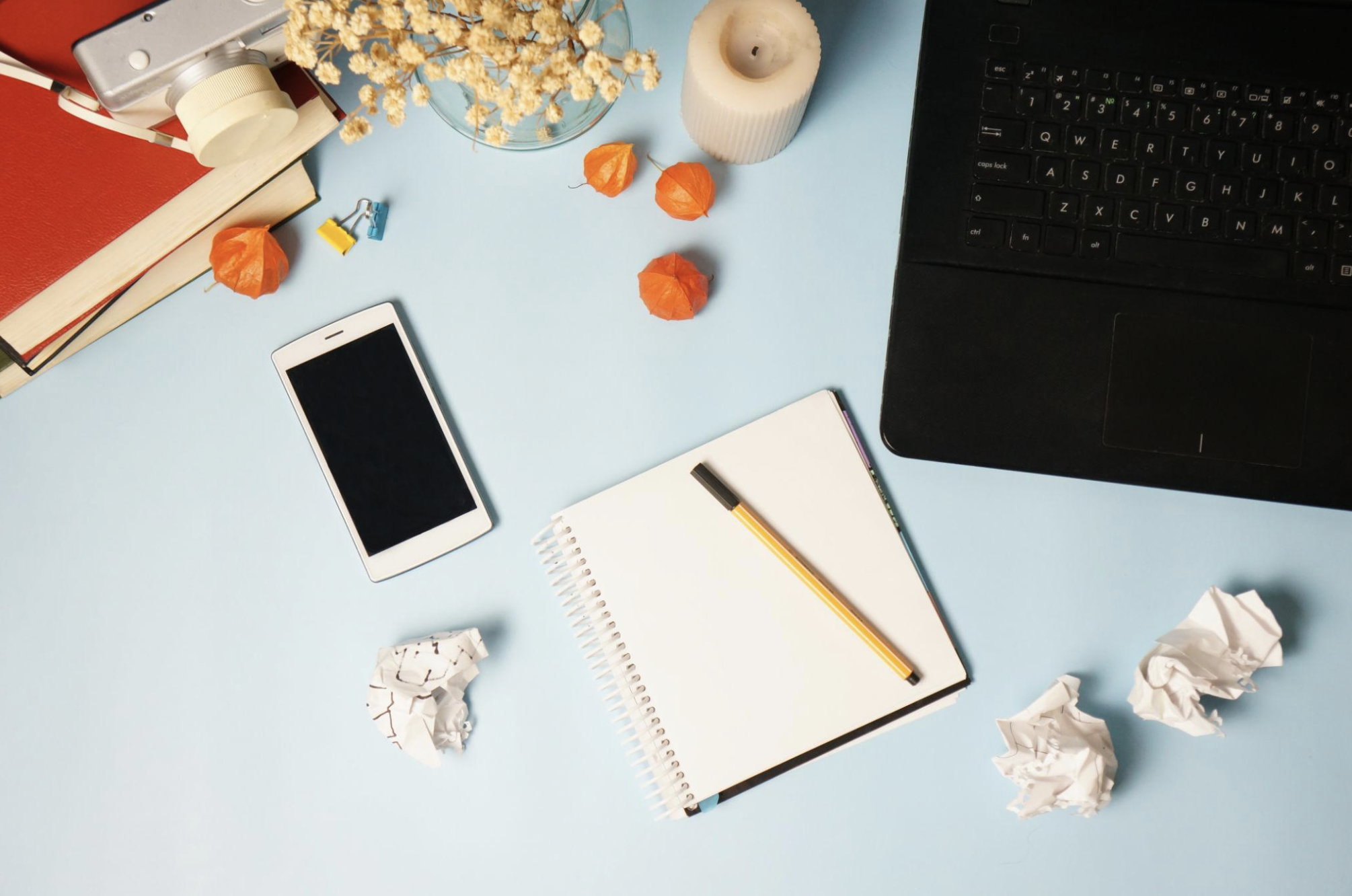 Overhead view of a cluttered blue desk with a black laptop, a white smartphone, a notepad with a yellow pen, a white candle, dried flowers in a glass bowl, crumpled papers, a red binder, and colored paper clips.