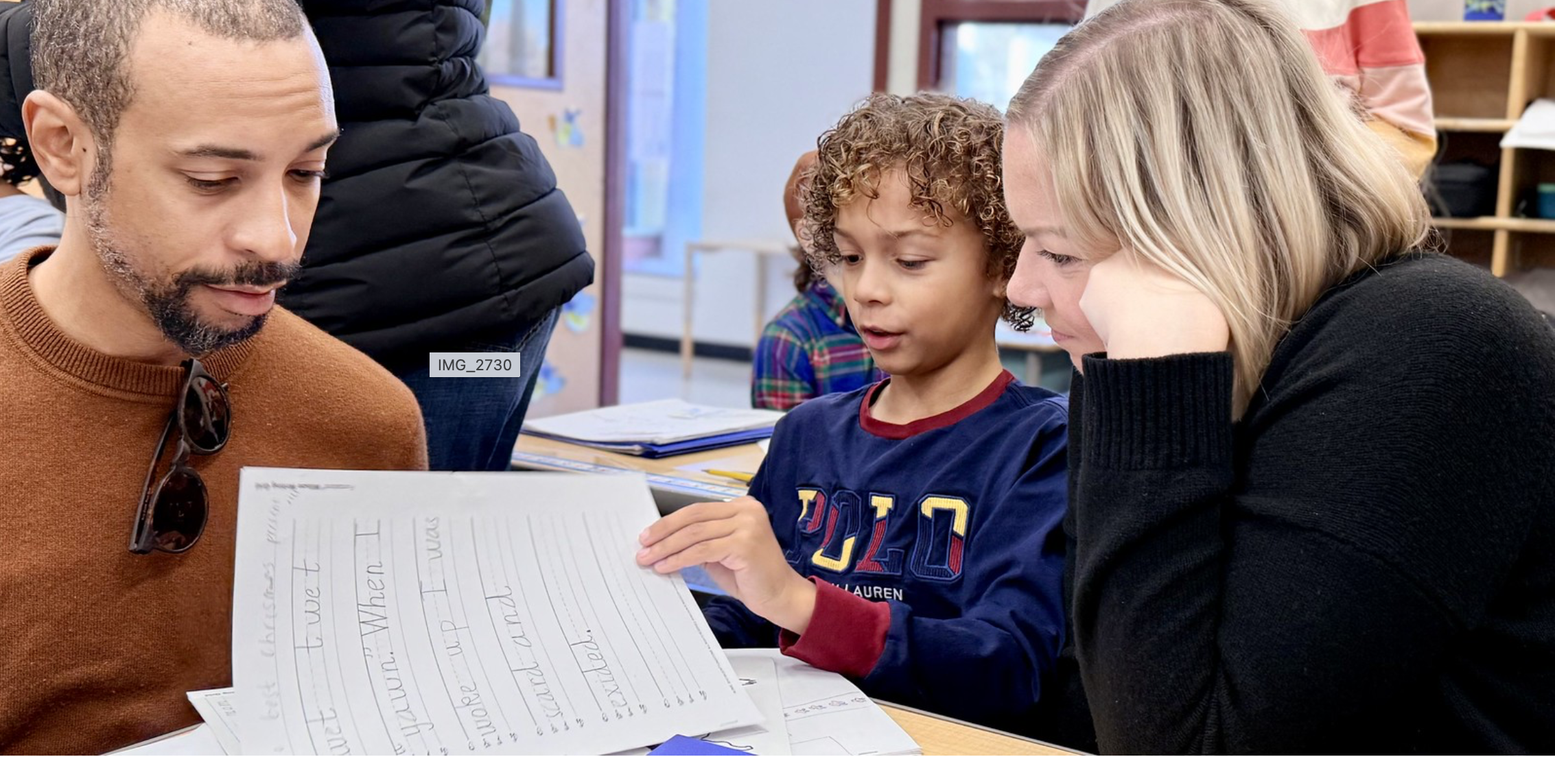 A man, a woman, and a young boy sitting together at a table looking at a sheet of paper with handwritten text. The setting appears to be a classroom or library, with bookshelves and windows in the background.