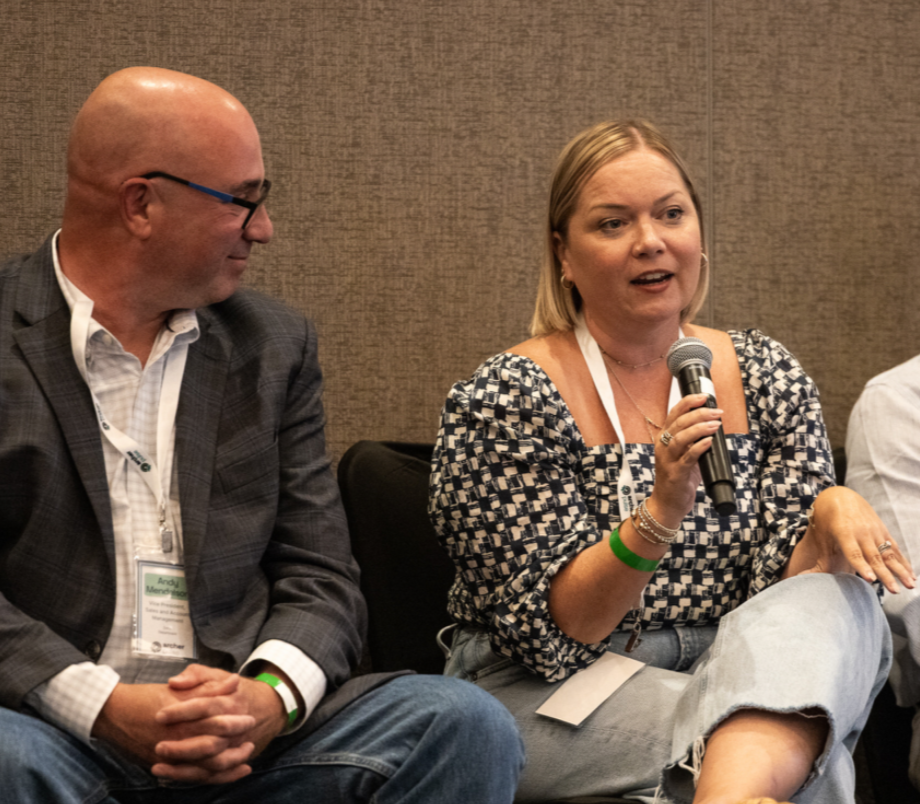 A woman speaking into a microphone at a conference, sitting next to a man in a suit who is listening