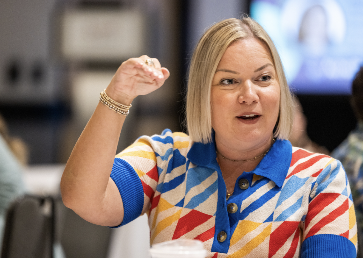 Woman with blonde hair wearing a multicolored geometric-patterned shirt, gesturing with her arm raised in a conversation or presentation, in an indoor setting.