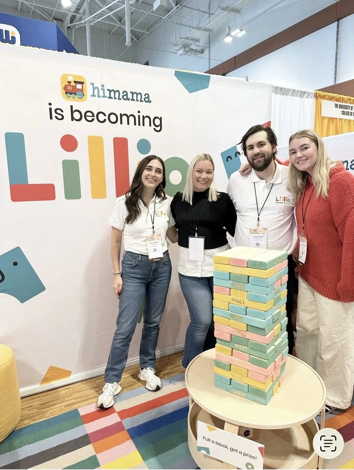 Four people standing behind a Jenga tower made of pastel-colored foam blocks at an event booth. The booth has a sign that reads 'Himama is becoming Lililo,' with colorful letters and floating game pieces in the background. A rainbow-patterned rug is on the floor, and a sign on the table says 'Pull a block, get a prize!'