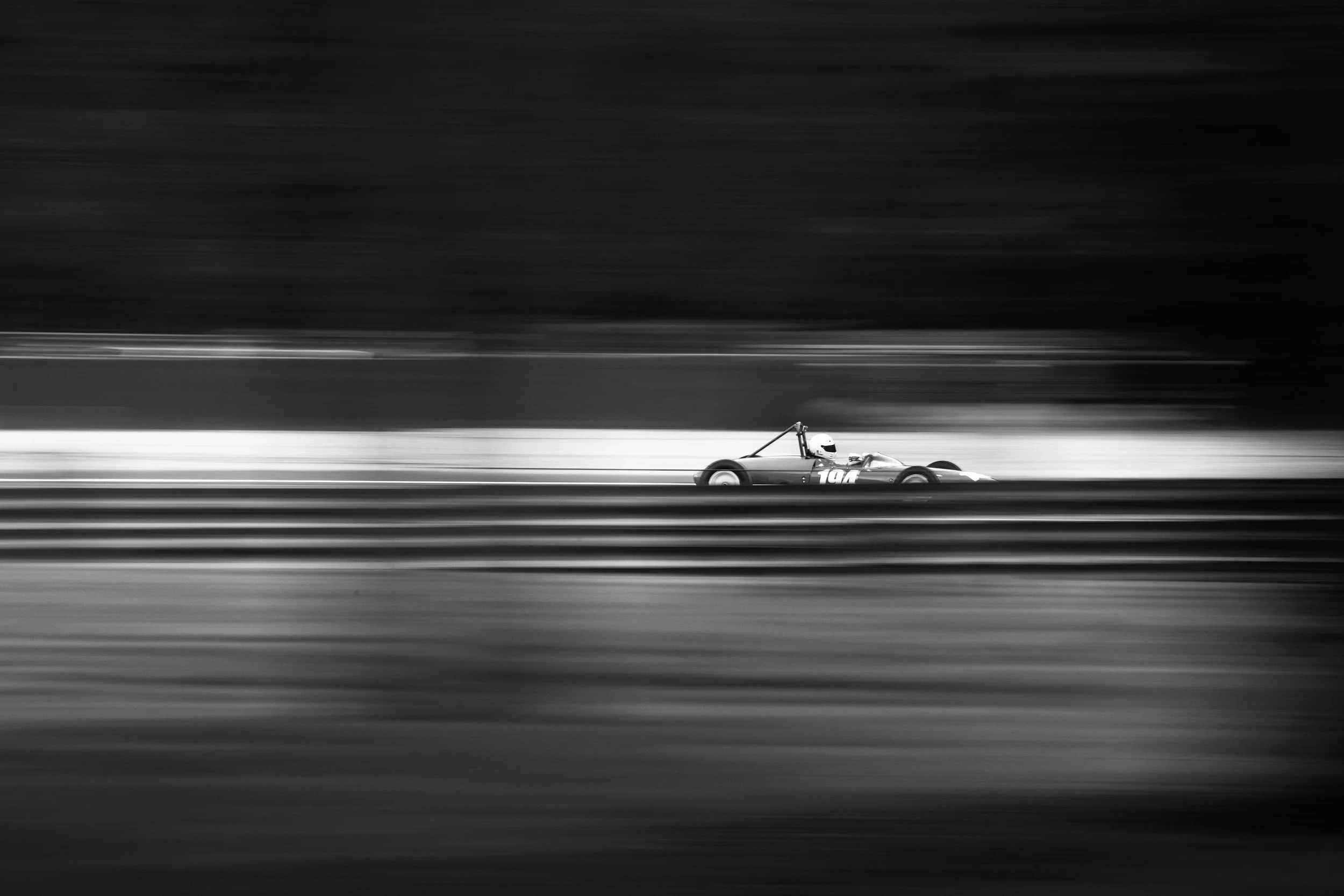 Black and white photograph of a race car moving at high speed on a track with blurred background and foreground, creating a sense of motion.
