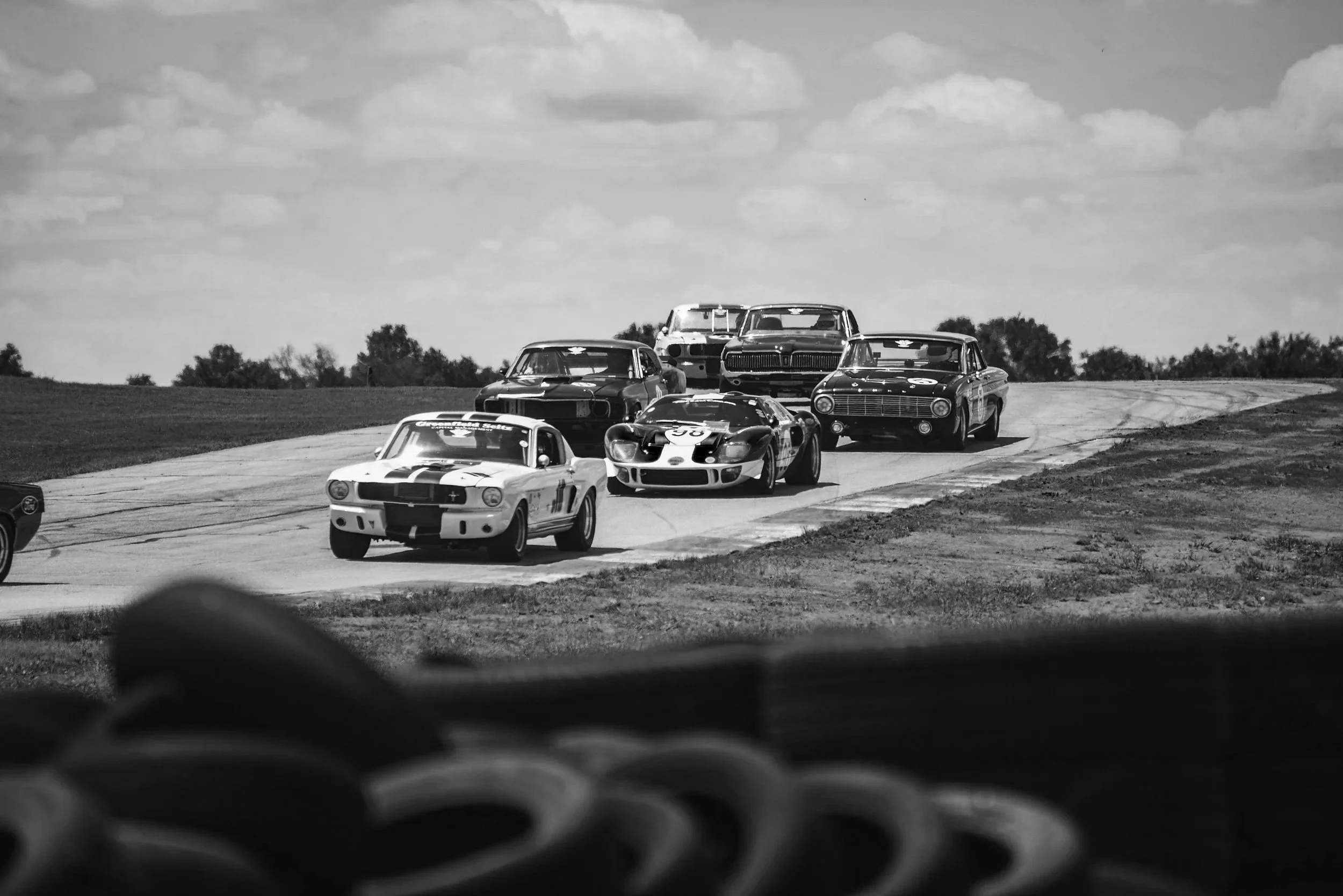 Black and white photo of vintage race cars on a race track, closely following each other in a competition, with the foreground slightly blurred.