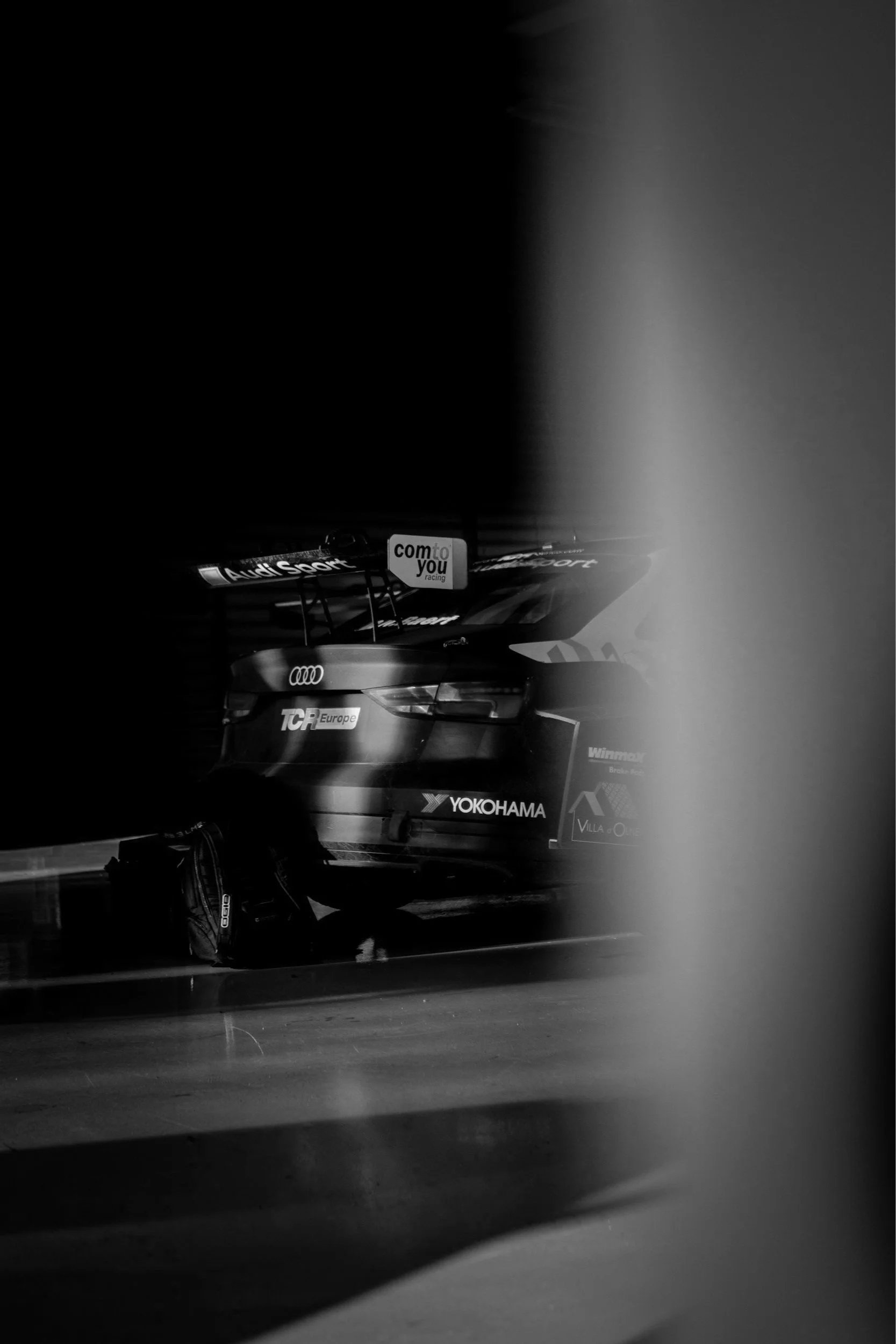 Black and white photo of a race car garage, showing the rear of an Audi race car with sponsor logos including Yokohama, TCR Europe, and com to you racing, partially obscured by a blurred column or barrier.