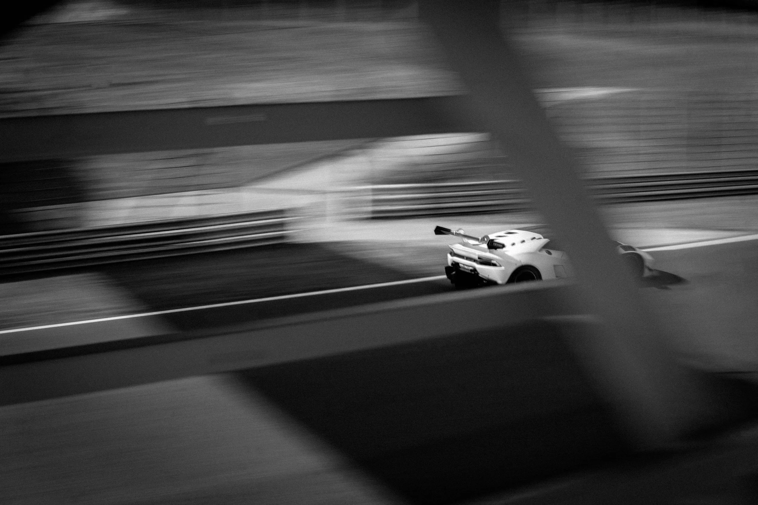 Black and white photograph of a race car on a track, viewed through a fence with blurred motion in the foreground.