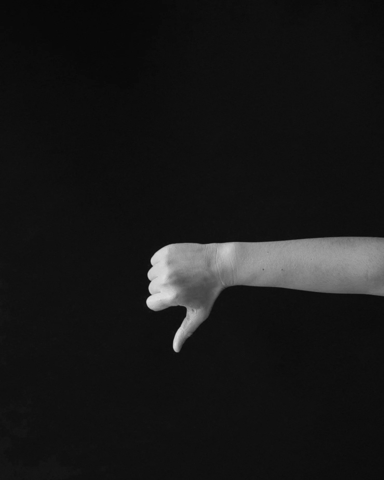 A black-and-white photo of an outstretched arm with a clenched fist, giving a thumbs-down gesture, against a dark background.