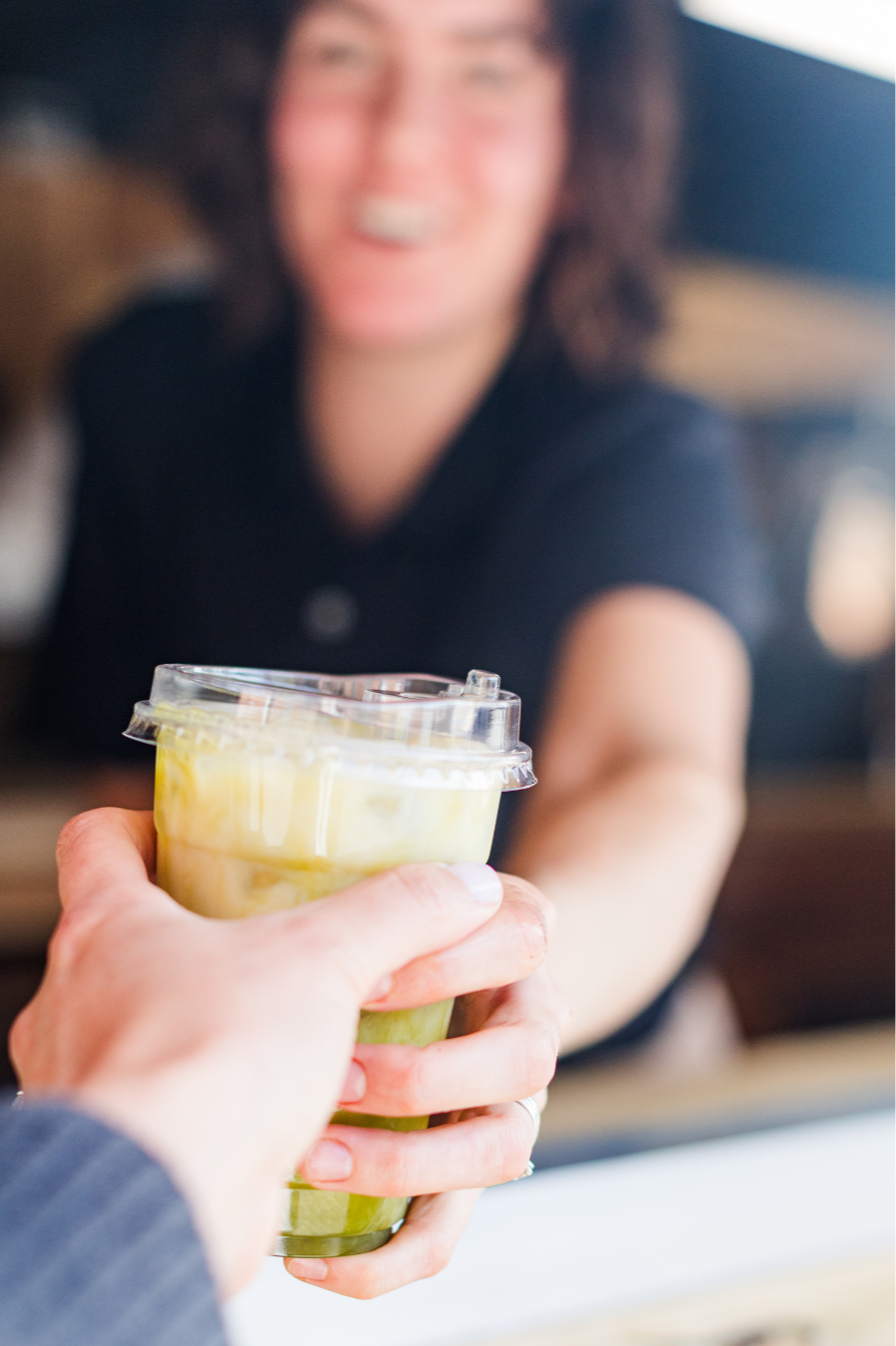 A person is handing a cup of green and yellow layered beverage to a smiling woman at a counter.
