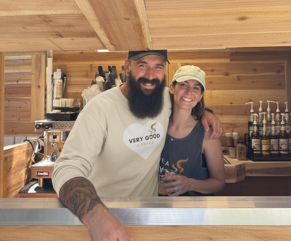 A man and woman smiling behind a wooden coffee bar in a coffee shop, with coffee bottles and equipment in the background.