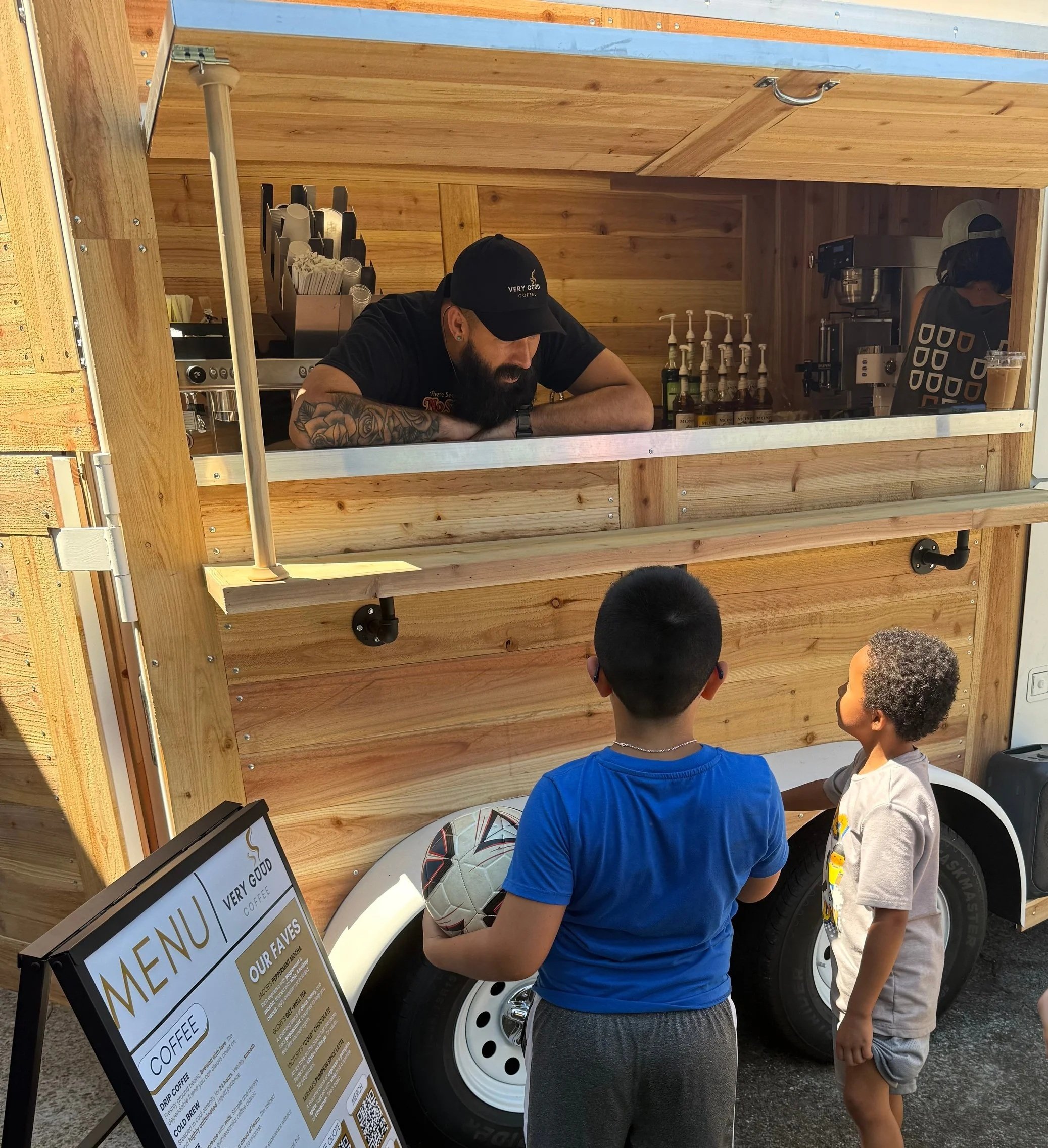 A man with a beard and tattooed arms, wearing a baseball cap and black T-shirt, stands behind a wooden food truck counter, holding a soccer ball. Two children with short hair, one with glasses, are standing in front of him, looking at the food truck menu. The food truck interior has a coffee machine, syrup bottles, and a menu sign with coffee options. The scene is outdoors, and the children are wearing casual clothing.