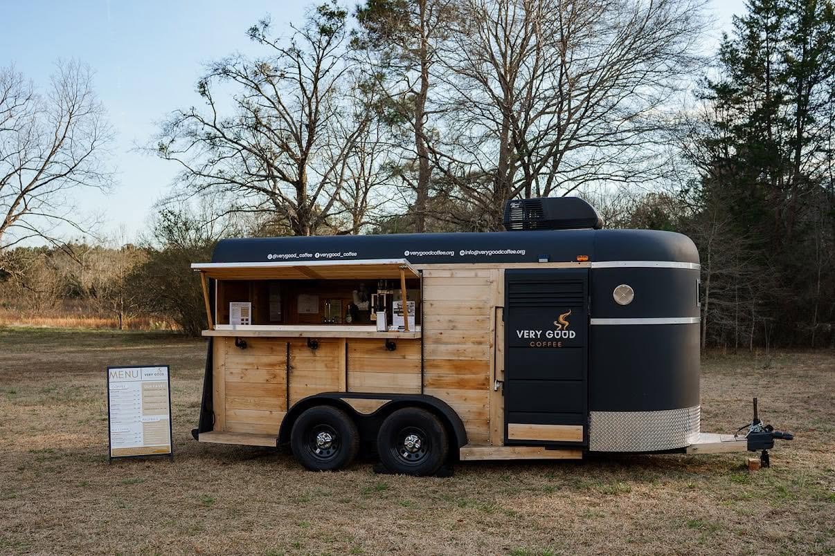 A black and wooden coffee trailer with a menu board in front, sitting on grassy field with leafless trees in the background.