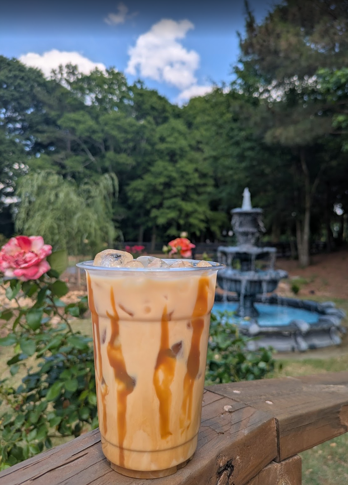 A glass of iced caramel coffee with ice cubes and caramel syrup drizzled on top, placed on a wooden railing outdoors with a fountain, trees, and flowers in the background.