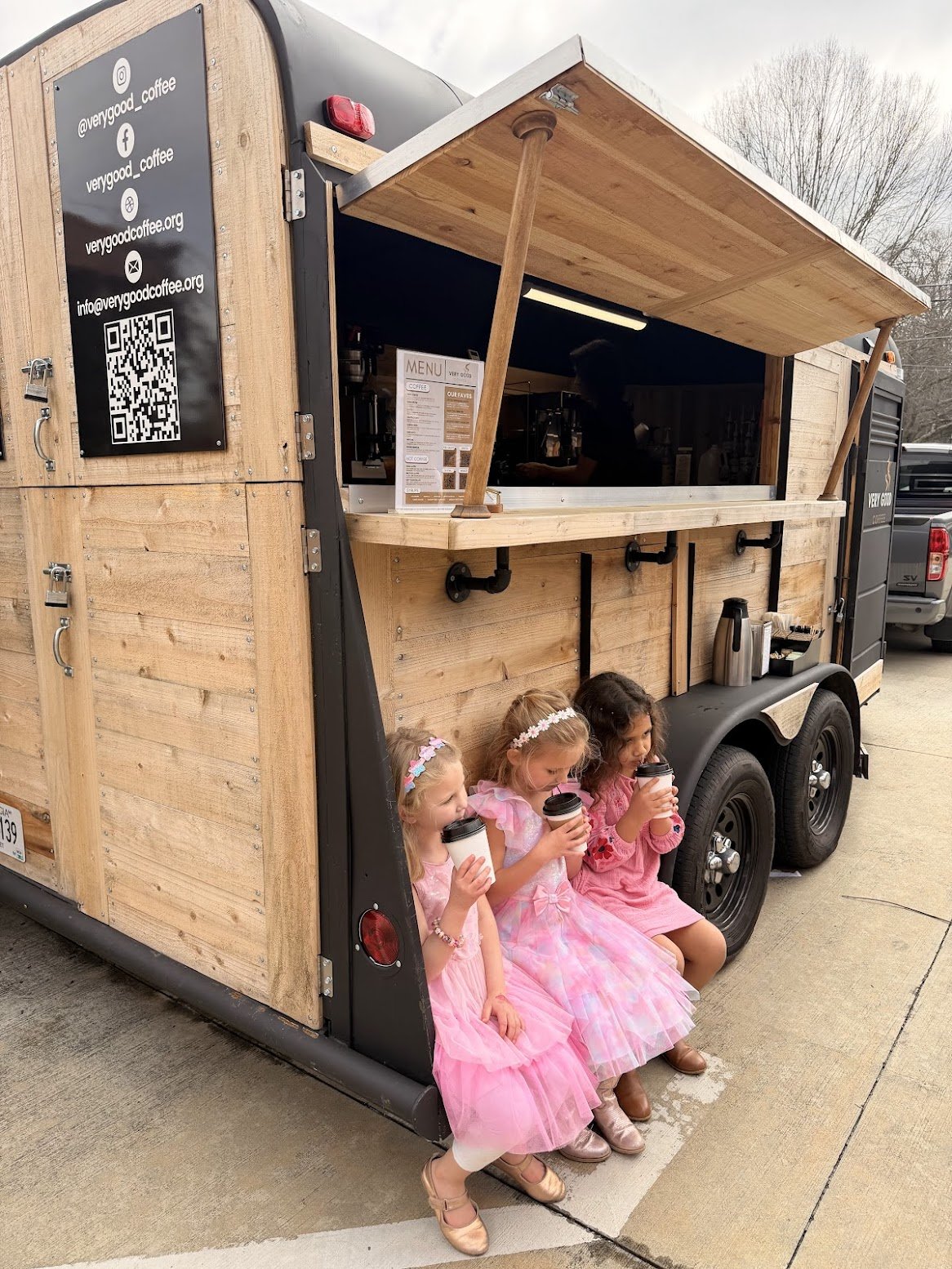 Three young girls in pink dresses sitting outside a food truck, enjoying beverages.