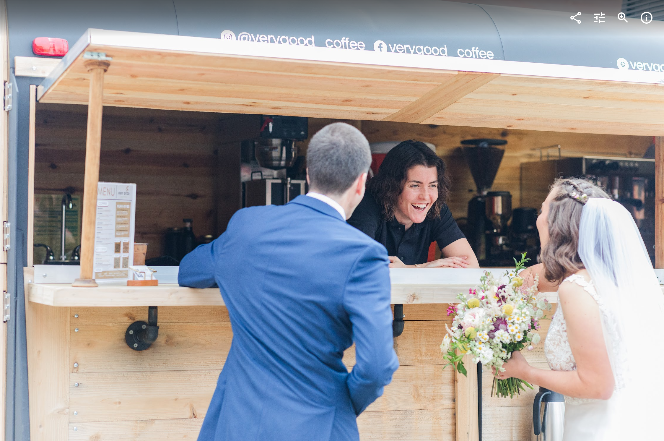 A woman in a wedding dress holding a bouquet of flowers talking to a man in a blue suit at a coffee stand, with a smiling female barista serving them.