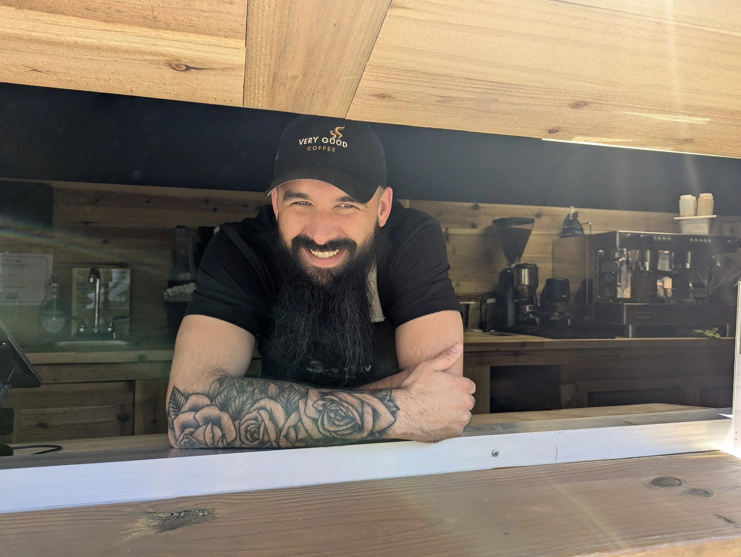 A smiling man with a beard and tattoos on his arm, wearing a black cap that says "Very Good Coffee," leaning on a counter inside a coffee shop.