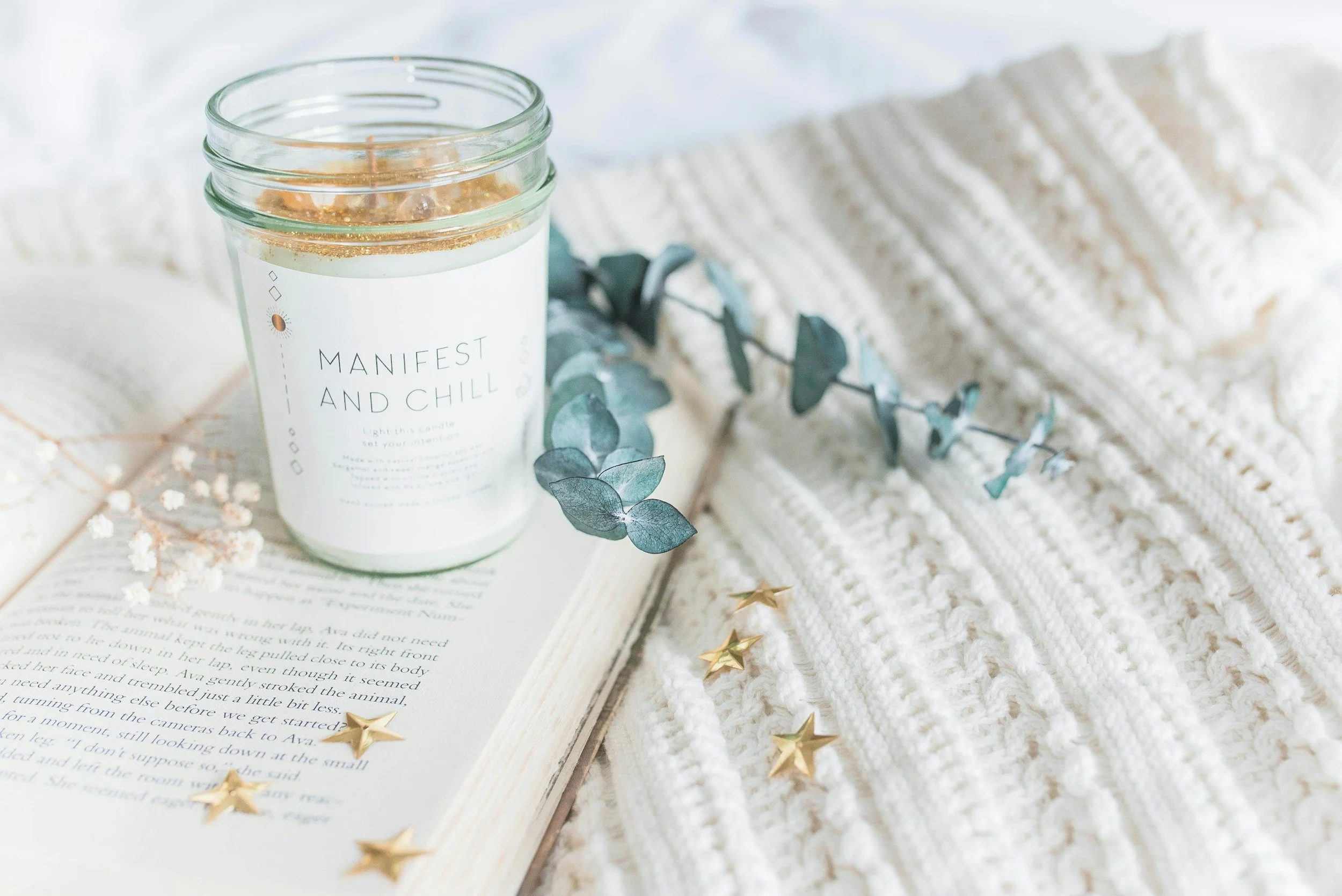 A lit white candle in a glass jar with a minimalist label reading 'Manifest and Chill', placed on an open book with dried flowers and eucalyptus leaves, surrounded by a plush white blanket and gold star confetti.