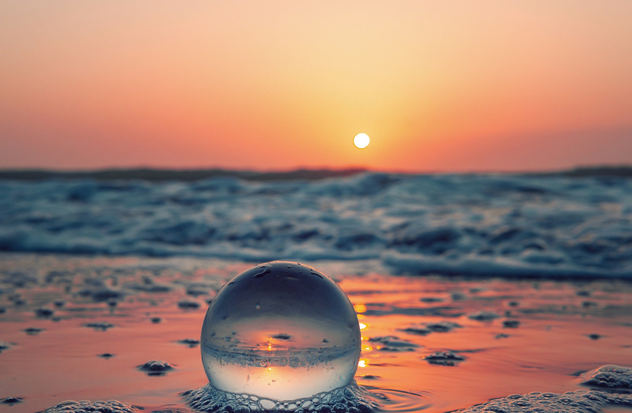 A glass ball on wet sand reflects the sunset, with a pink and orange sky and the sun low on the horizon over the ocean waves.