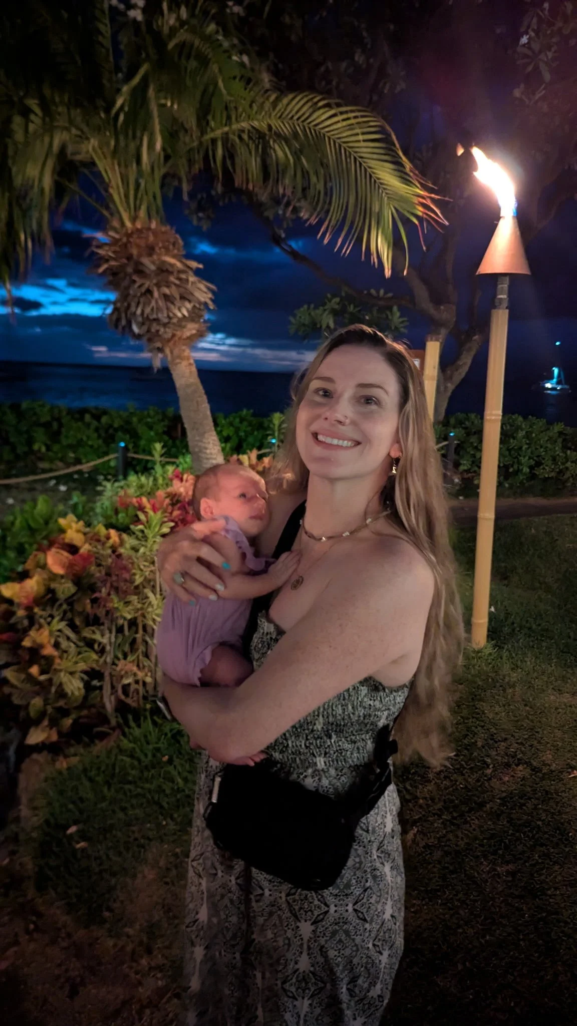 A woman holding a newborn baby outdoors at night near a tiki torch, palm trees, and tropical plants, with a dark ocean and a partly cloudy sky in the background.