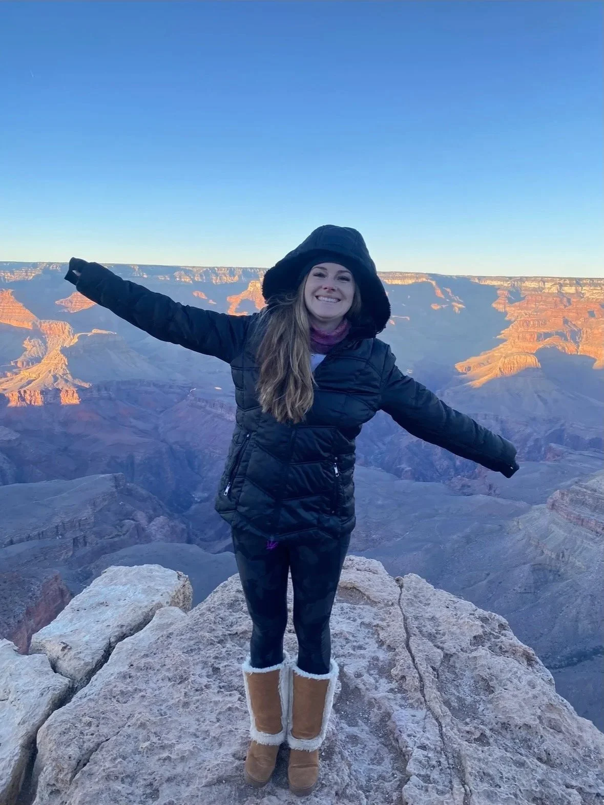 A woman smiling with arms outstretched standing on a rocky ledge at the Grand Canyon during sunset.