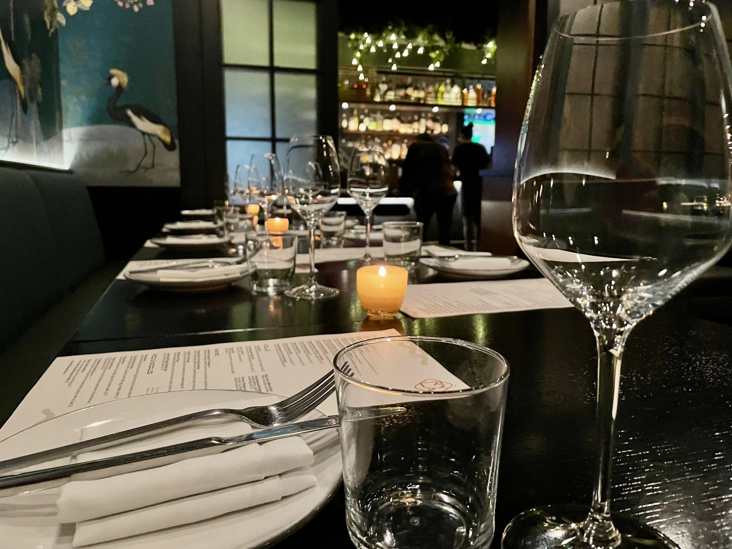 Elegant restaurant table setting with wine glasses, plates, cutlery, and menus, featuring lit candles and dim ambient lighting, with a bar visible in the background.