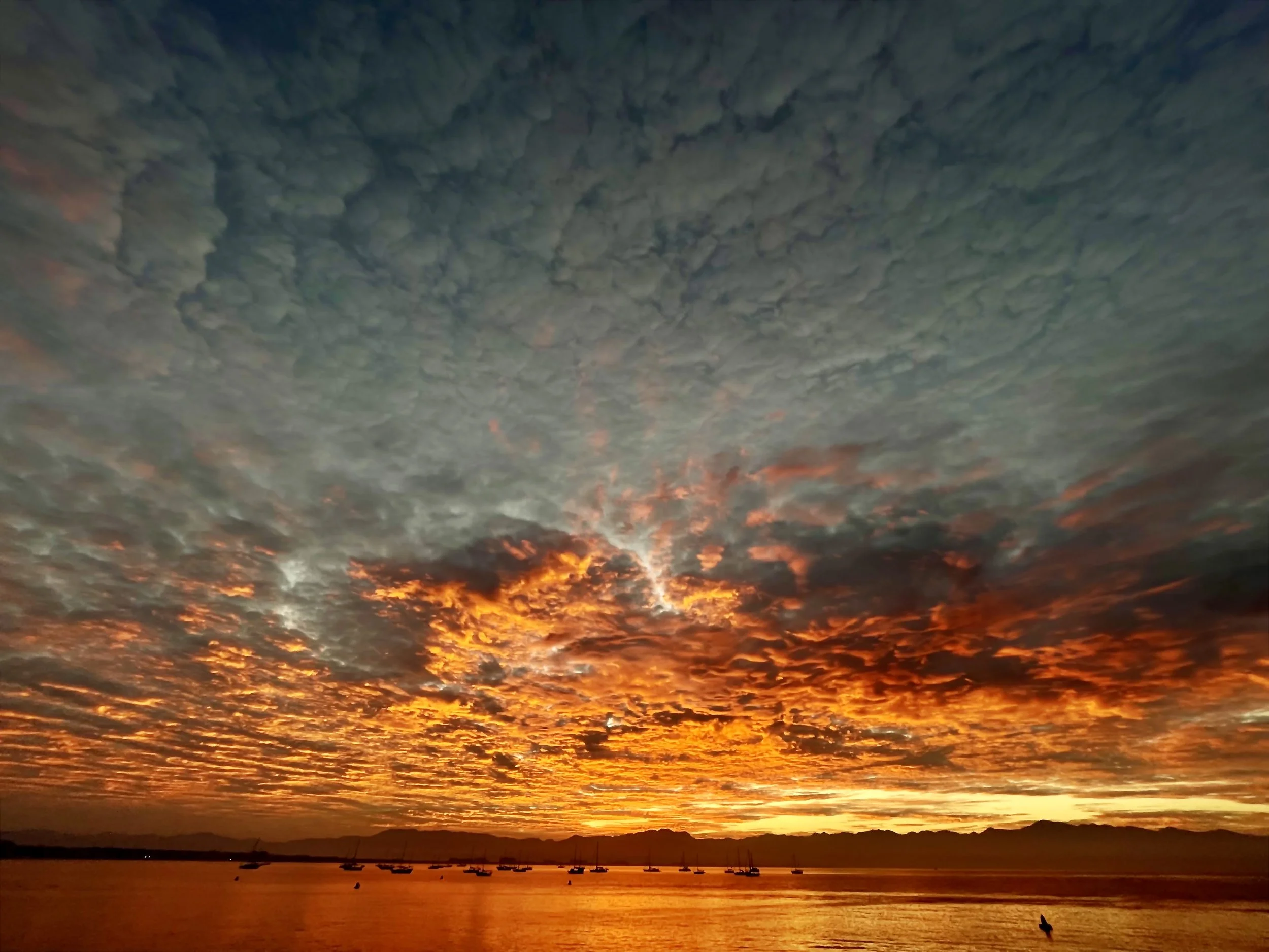 Dramatic sunset over a calm sea with vibrant orange and dark clouds, silhouetted boats on the horizon.