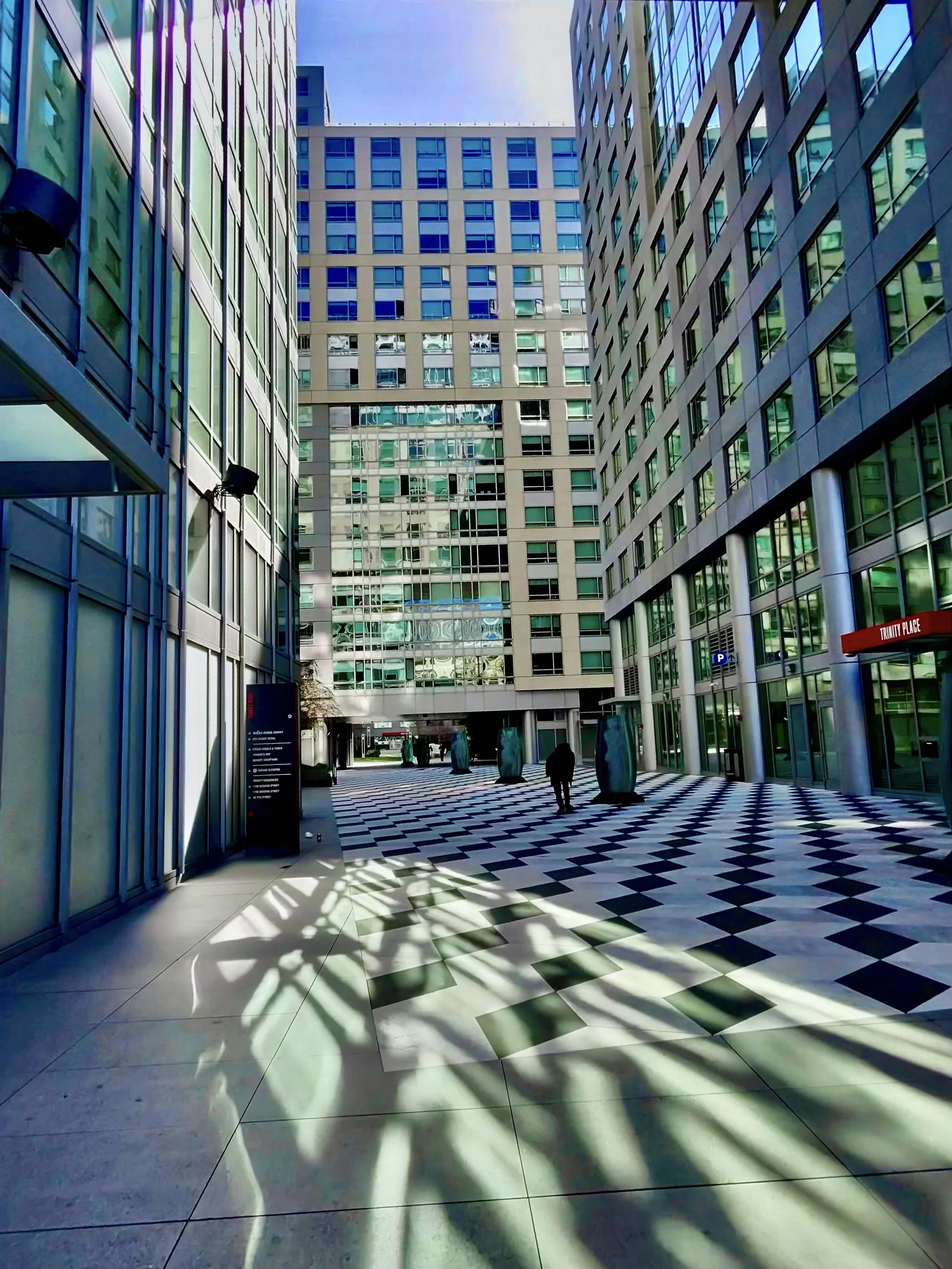 Outdoor urban area with tall glass buildings, checkered floor, shadows, and a person walking away.