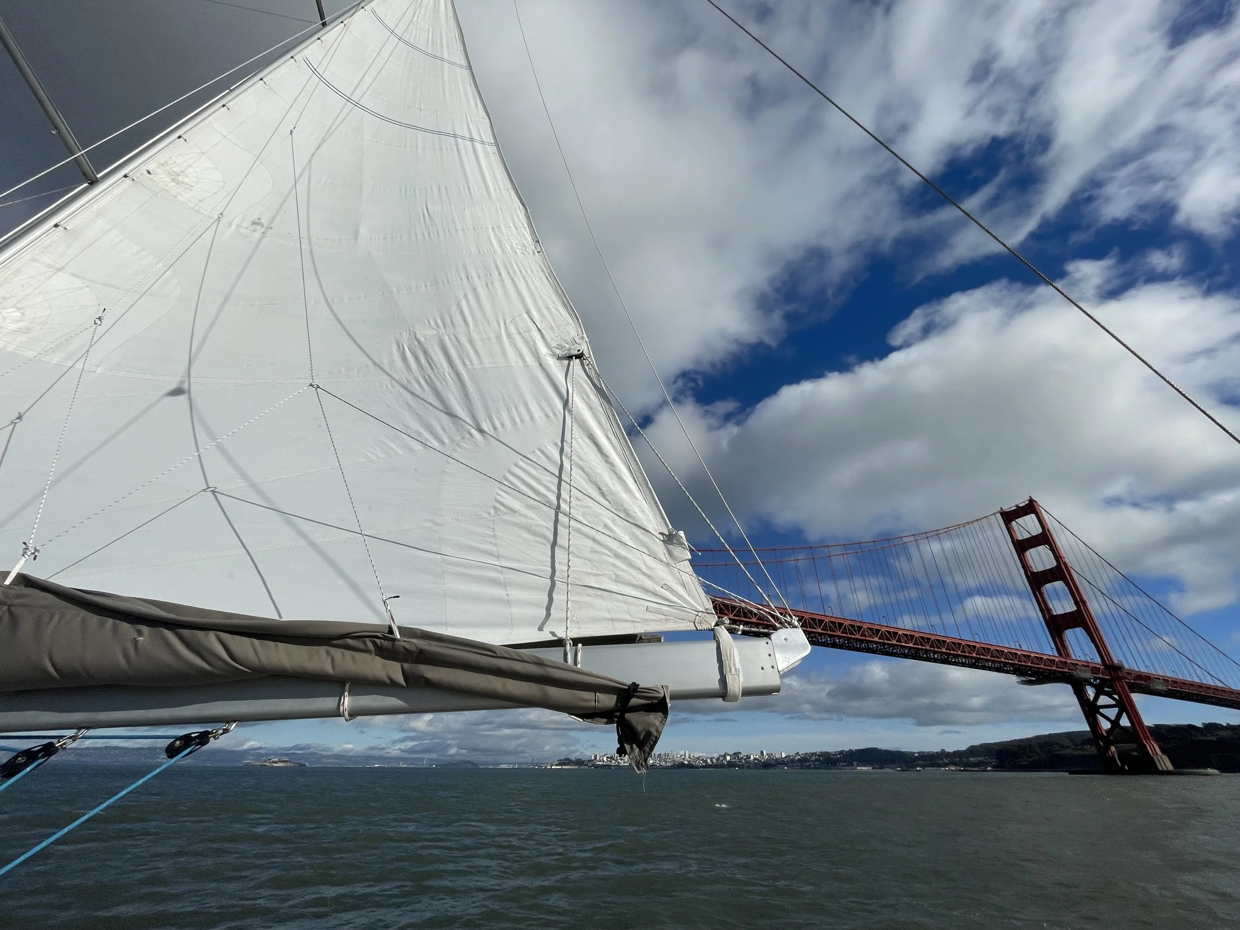 View of a sailboat sail with the Golden Gate Bridge in the background, under a partly cloudy sky.