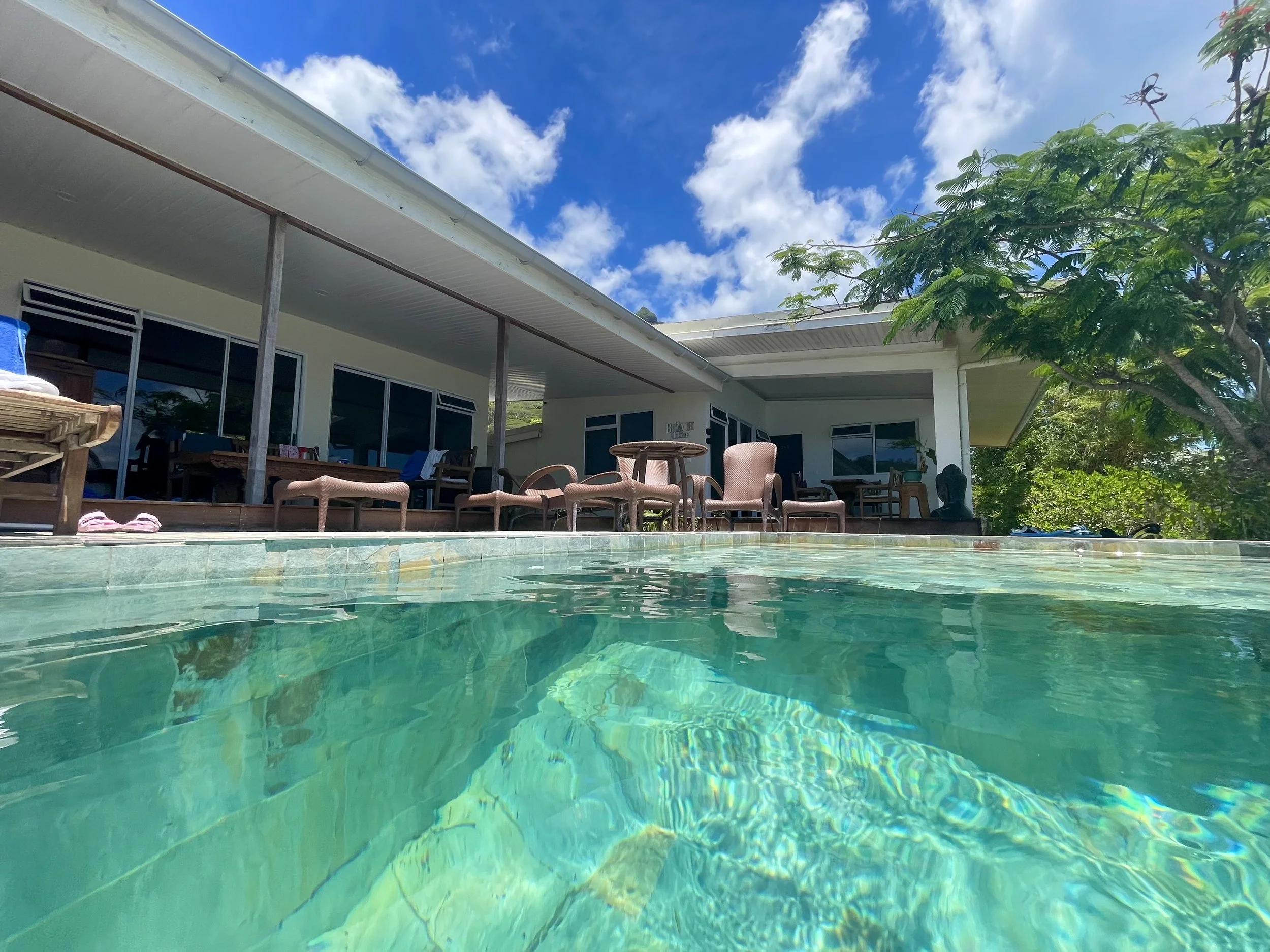 View of a luxurious outdoor pool next to a modern house with patio furniture under a covered porch, surrounded by lush greenery on a sunny day.